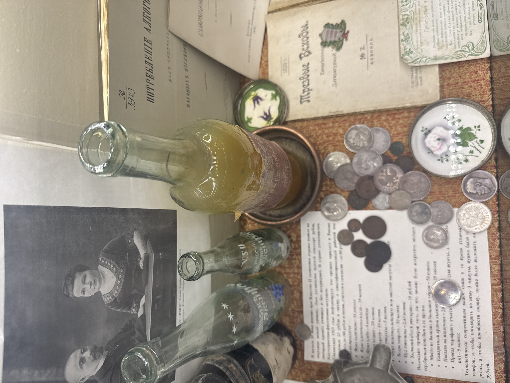 This image depicts a collection of vintage items, likely displayed in a museum or historical exhibit, possibly in Saint Petersburg, Russia, given the Cyrillic text and the nature of the objects.
The items are arranged on a surface with a patterned fabric. Several glass bottles are visible, one containing a yellowish liquid. There is an assortment of old coins. Several small, decorative dishes or boxes with floral designs are present. Open books and documents written in Cyrillic are scattered around. One document is titled "ПОТРЕБЛЕНИЕ АЛКОГОЛЯ" (Consumption of Alcohol). There is also a black and white photograph of a woman and a man. One of the bottles has Cyrillic markings and the word "Русское" which translates to "Russian." Other documents appear to list prices for various goods and services. One document is titled "Трезвые Всходы" which could translate to "Sober Sprouts."