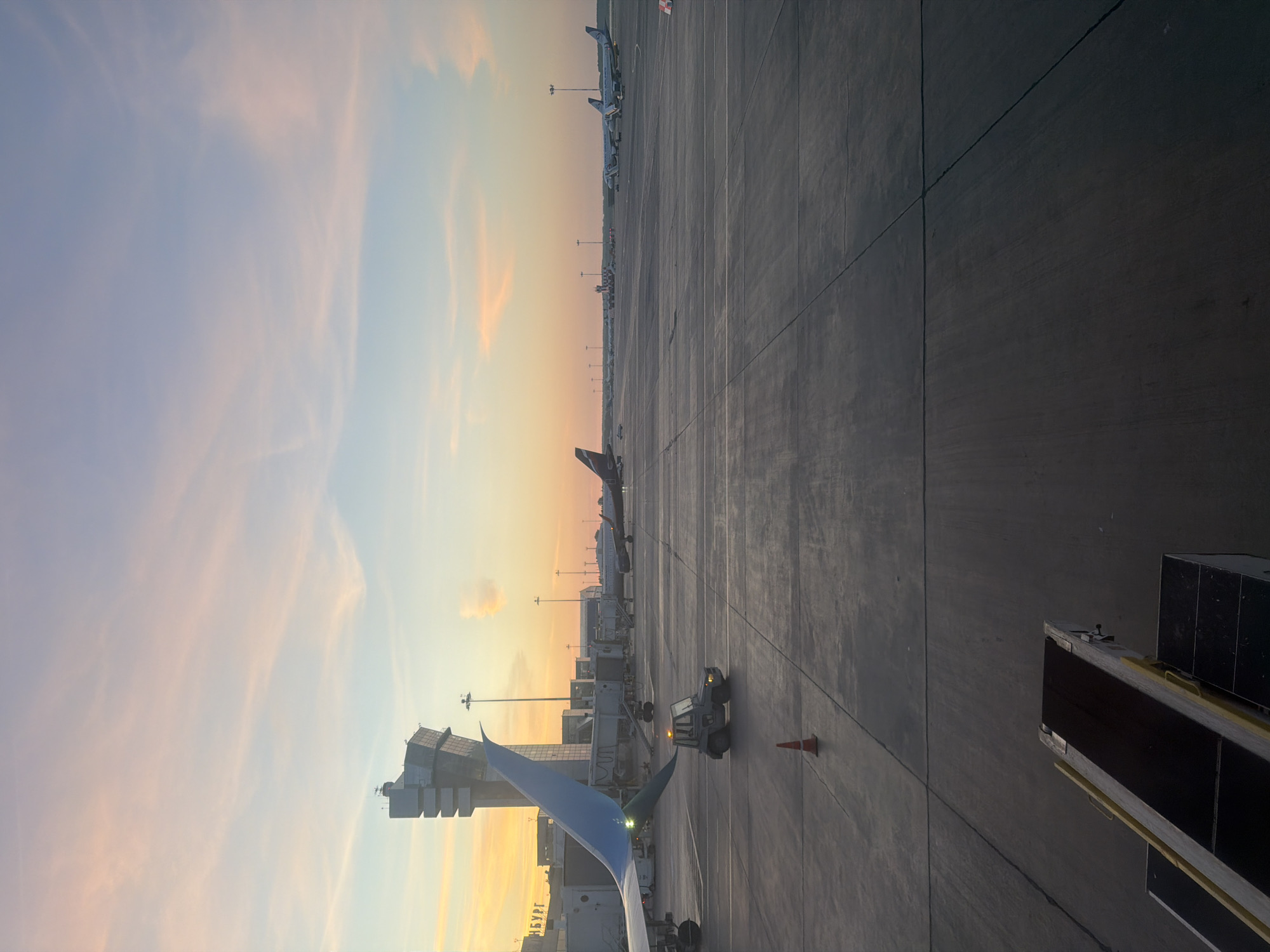 The image depicts an airport tarmac at sunrise or sunset, with soft pastel colors in the sky. In the foreground, part of a baggage conveyor belt is visible. To the left, an aircraft wing with a blue tip is prominent, attached to a jet bridge. A small tug vehicle is positioned on the tarmac near a bright orange traffic cone. Several airplanes are parked on the tarmac in the middle and background, along with airport buildings and a control tower. The control tower has a distinct architectural style with several stacked rectangular sections. The overall scene suggests a quiet moment at an airport before or after operations, bathed in the warm glow of the low sun. The presence of Russian text on the building in the background ("НБУРГ") indicates the location is likely Pulkovo Airport in Saint Petersburg, Russia, as it is a common abbreviation for the city.