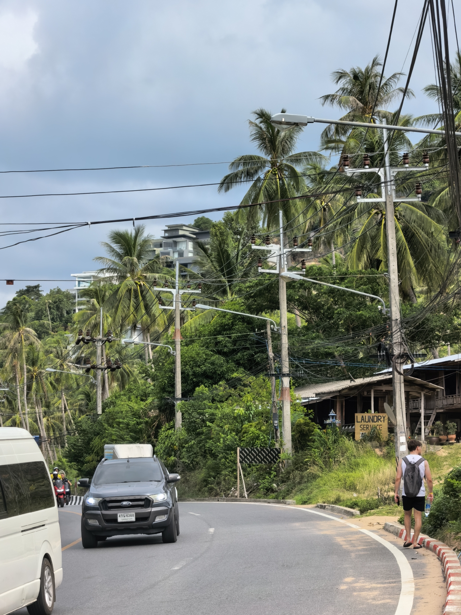 The image depicts a street scene in the Ko Samui District of Thailand, characterized by lush tropical vegetation, palm trees, and overcast skies. In the foreground, a gray Ford Ranger pickup truck is driving on a paved road, with its headlights on. To the left, the rear of a white van is visible. Further down the road, two motorcycles are seen. A man wearing a white tank top, black shorts, a backpack, and flip-flops is walking away from the camera on the sidewalk, holding a water bottle. Beside him, a yellow sign reads "LAUNDRY SERVICE." The road is lined with utility poles carrying numerous electrical wires. In the background, a modern multi-story building is visible on a hillside, surrounded by dense green foliage and more palm trees. Several wooden structures are also present along the side of the road.