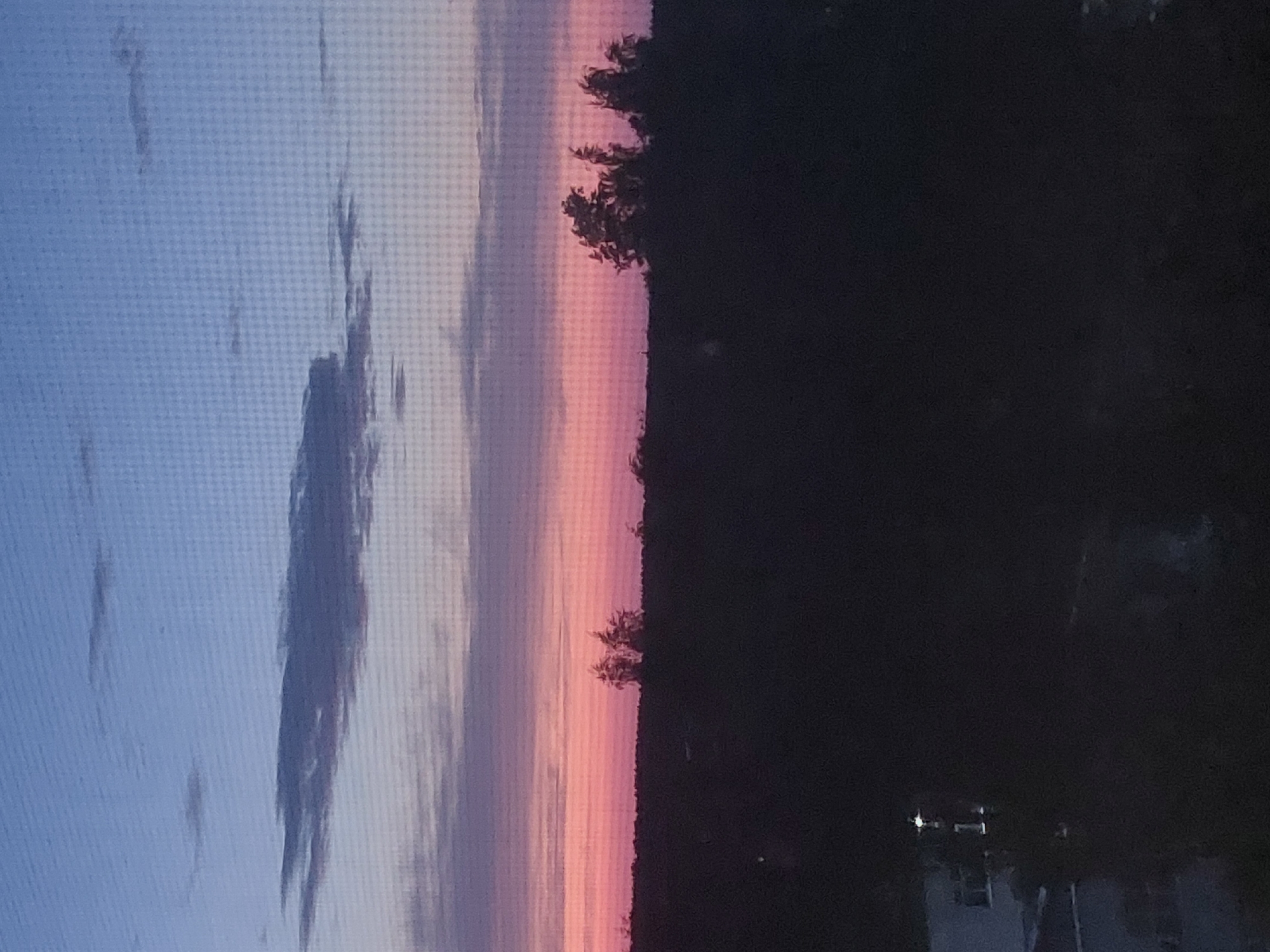 The image depicts a sunset scene with a clear division between the sky and land. The sky transitions from a blue with elongated clouds on the left, to pink and purple hues towards the center. A dark silhouette of trees stretches along the horizon, separating the colorful sky from the dark land area on the right side. In the dark land area, there are faint lights from a building. The location is Chaikovsky, Russia.