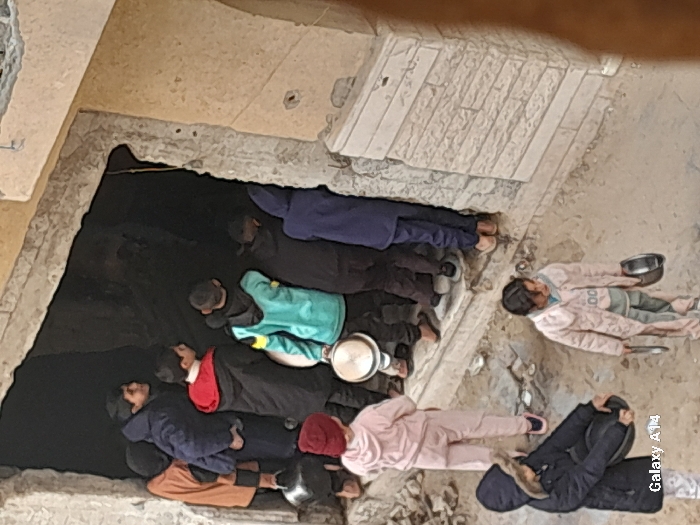 The image shows approximately nine children and young individuals gathered at and around a dark, rectangular entrance to a building in Khan Yunis, Palestinian Territory. Six individuals are positioned at the entrance, with several looking towards the interior or to the side. At least five individuals, both at the entrance and in the foreground, are holding metallic pots or bowls. They are dressed in various layered clothing, including jackets and hooded garments. The building's exterior features light-colored, possibly damaged, walls with visible concrete and rough edges around the doorway. The ground in the foreground is uneven dirt or rubble.
