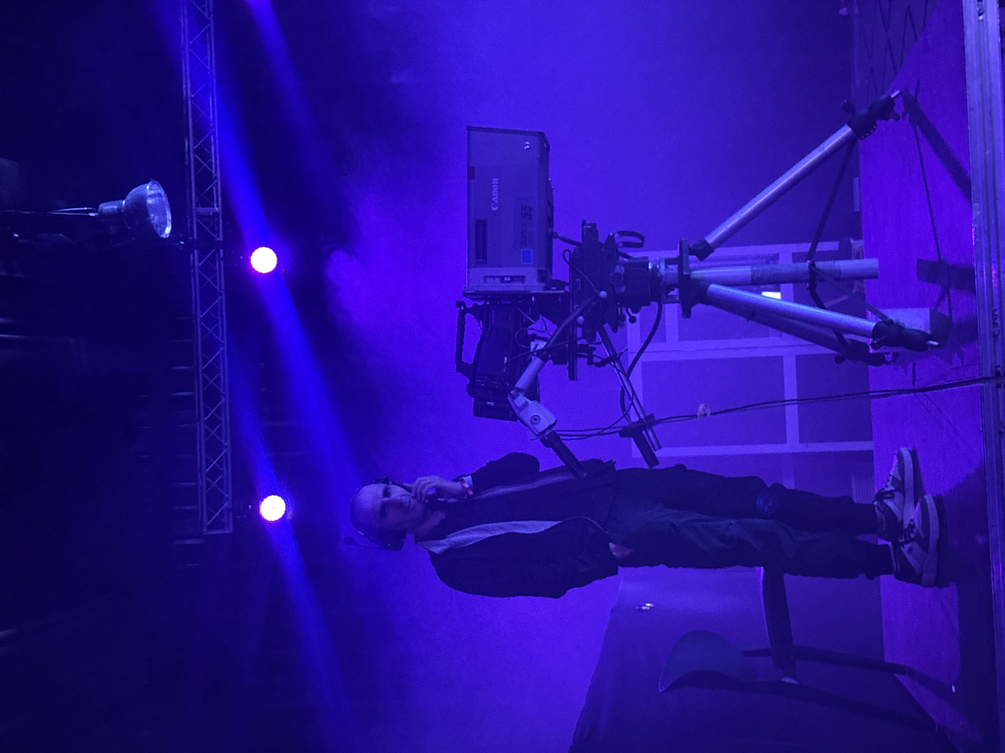 A male figure with a shaved head, wearing a headset and a dark jacket with a light stripe, stands next to a large Canon camera mounted on a tripod. The scene is illuminated with blue and purple lighting, suggesting a stage or broadcast setting. In the background, a light fixture and a support structure are visible. The location is not possible to determine with the image.