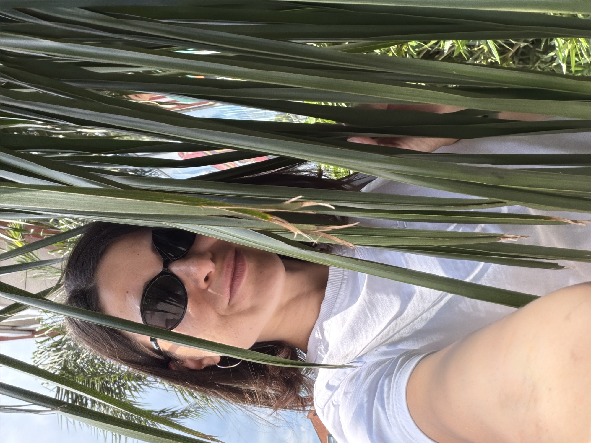 A woman with dark hair and sunglasses is visible through the fronds of a palm tree. She is wearing a white t-shirt and a silver hoop earring in her left ear. The background shows a building with a red roof and green foliage, and a glimpse of blue sky. This is located in Budva, Montenegro.