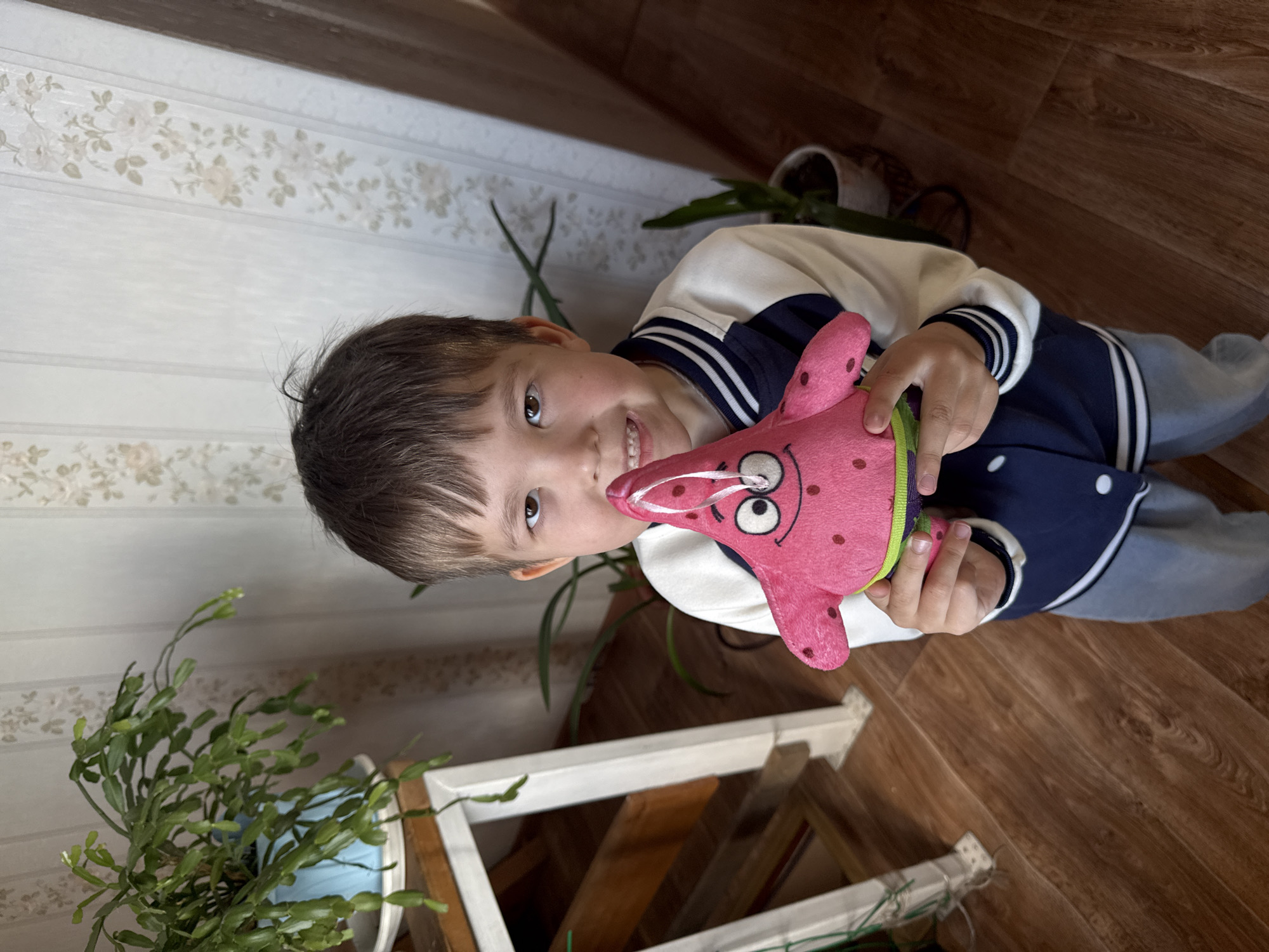 A boy is holding a Patrick Star plush toy. The location appears to be inside a home in Bely Yar, Russia, given the context. The boy has brown hair and is wearing a jacket with white and navy blue stripes. The background shows floral wallpaper, a plant, and wood flooring. A white wooden structure, possibly a plant stand, is also visible.