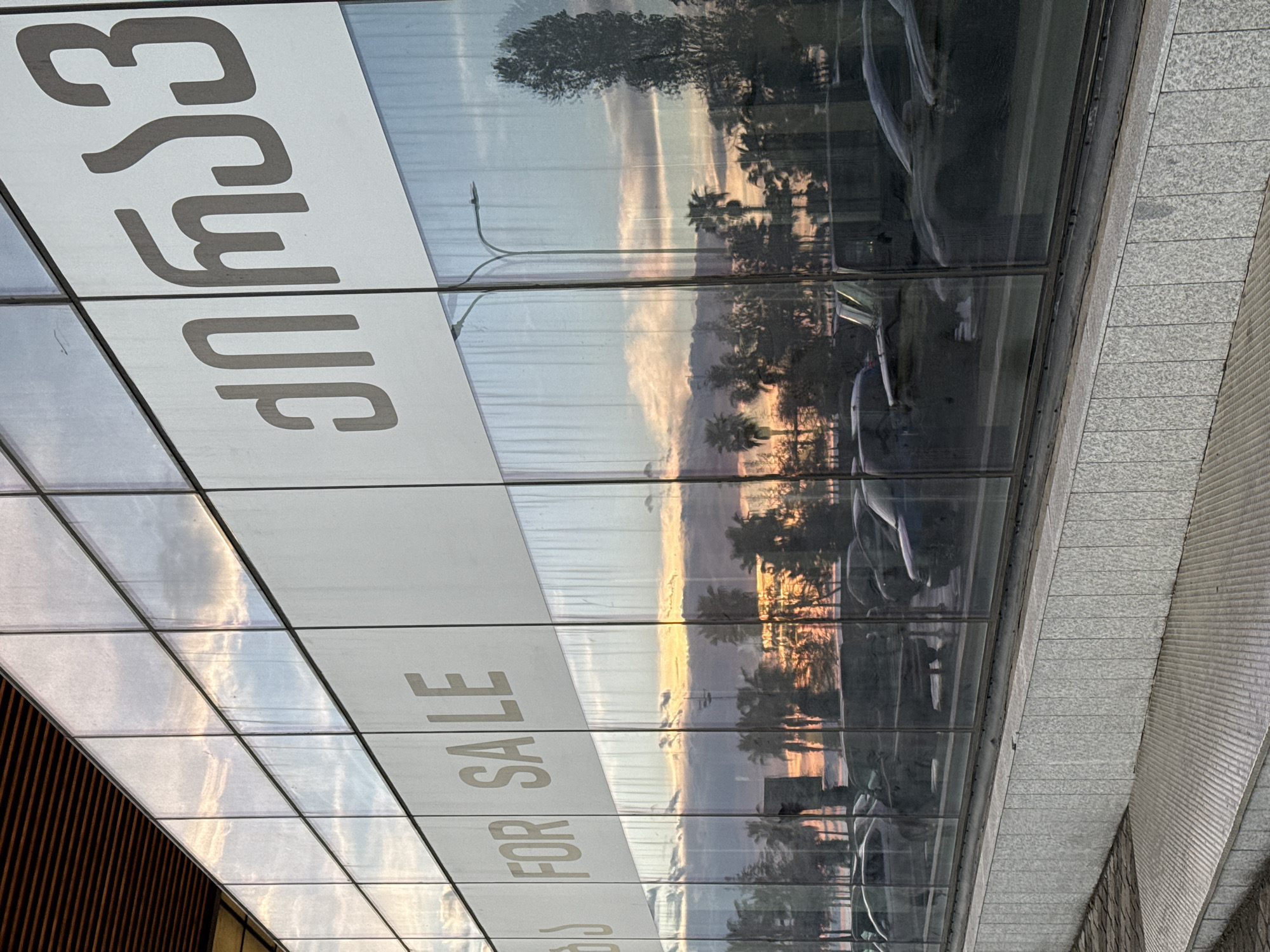 Here is a description of the image:
The image shows a reflective glass exterior, possibly a building facade. Bold, white lettering reading "იყიდება" (which translates to "For Sale" in Georgian) is visible on some of the glass panels. Below this, more white lettering states "FOR SALE" in English. The glass reflects a scene of trees and a sunset or sunrise sky. Cars are also faintly visible in the reflections. The facade is part of a building featuring concrete and a warm, wood-like material on parts of its structure. Given the Georgian text and the city's popularity for real estate, this scene could be located in Batumi, Georgia.