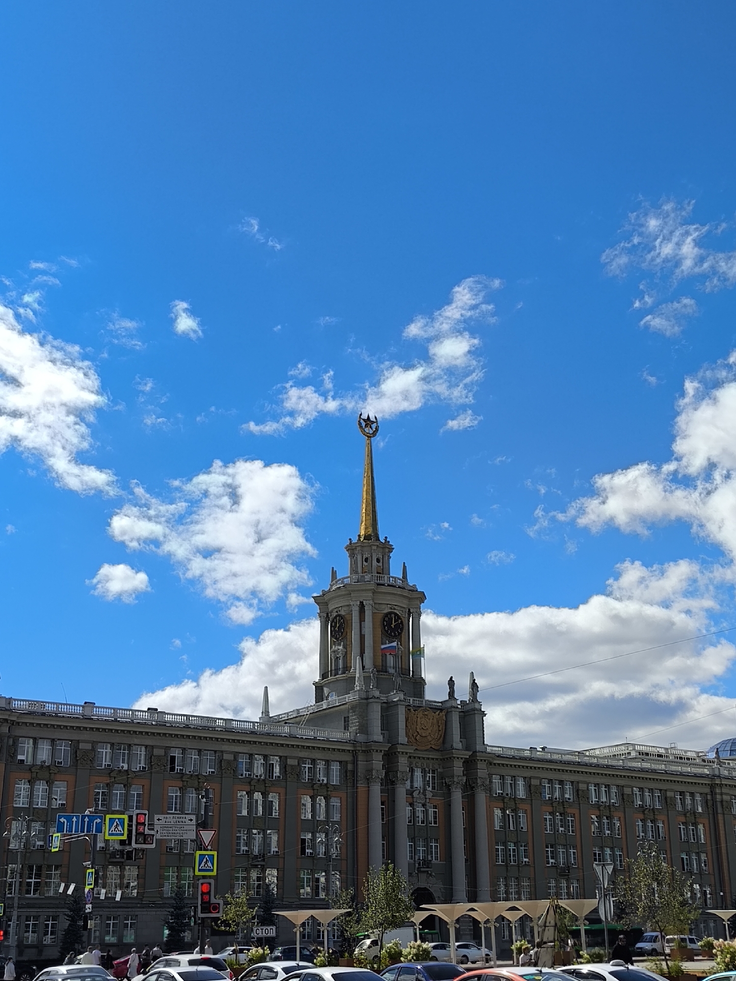 The image shows a daytime exterior view of the Yekaterinburg City Administration building, a prominent landmark in Yekaterinburg, Russia. The building is a large, multi-story structure with a central tower topped by a golden spire featuring a star. The architecture is classical with columns and decorative elements. A clock face is visible on the tower. The sky above is bright blue with scattered white clouds. In front of the building, there is a street with several cars, traffic lights, and pedestrian crossings, indicating an urban environment. Pedestrians are also visible on the sidewalk and near the crosswalks. The overall scene depicts a bustling city day in front of a significant administrative building.