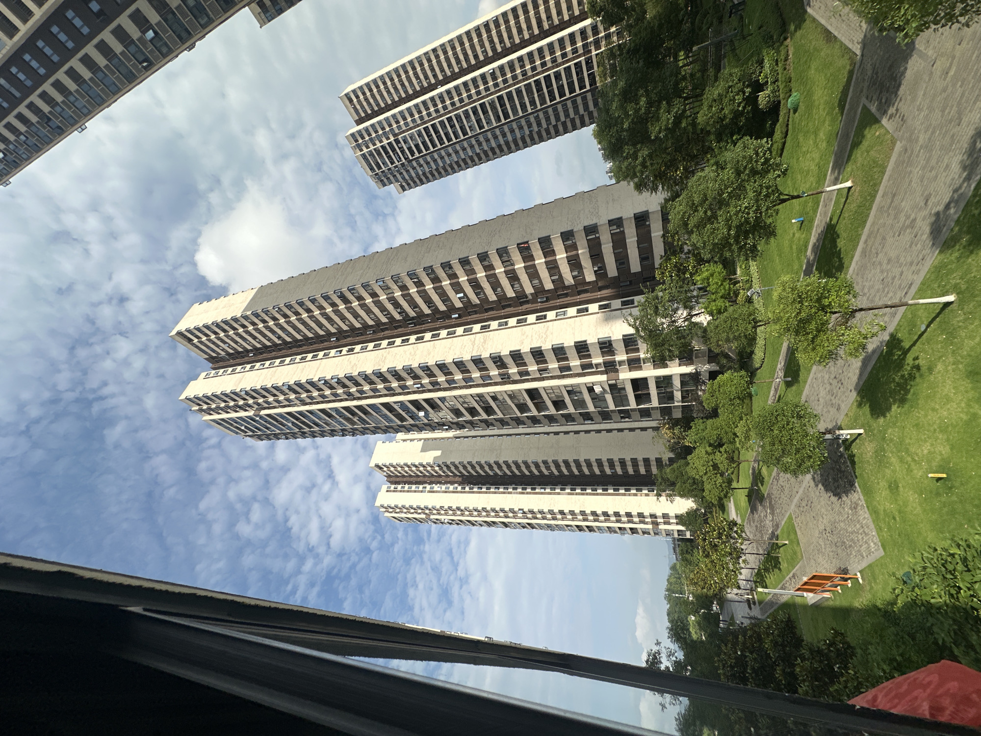 The image, taken from an elevated position in Chongqing High-tech Zone, China, showcases a cluster of high-rise apartment buildings against a partially cloudy sky. The buildings are light-colored with dark window accents and stand adjacent to a landscaped green space featuring trees, grass, and paved walkways. The bottom portion of the frame includes part of a window, which provides a view of the buildings.