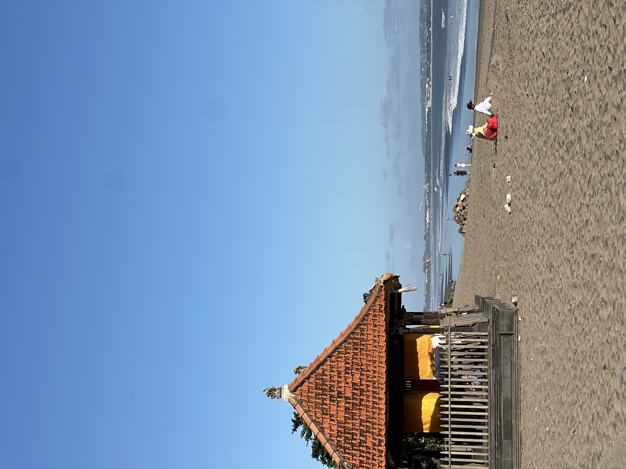 The image depicts a wide, sandy beach in North Kuta, Indonesia, under a clear blue sky. In the foreground, to the left, is a small, traditional Balinese structure with an orange tiled roof and yellow walls, featuring a wooden fence. Further into the scene, a body of water with moderate waves meets the sandy shore. Several individuals are visible on the beach and in the water. On the sand, a group of people are seated, appearing to relax or converse. In the distance, a city skyline is discernible, with buildings and structures stretching along the coast. An airplane is visible in the sky, suggesting proximity to an airport. The overall scene conveys a serene beach environment with signs of human activity and a glimpse of urban life in the background.
