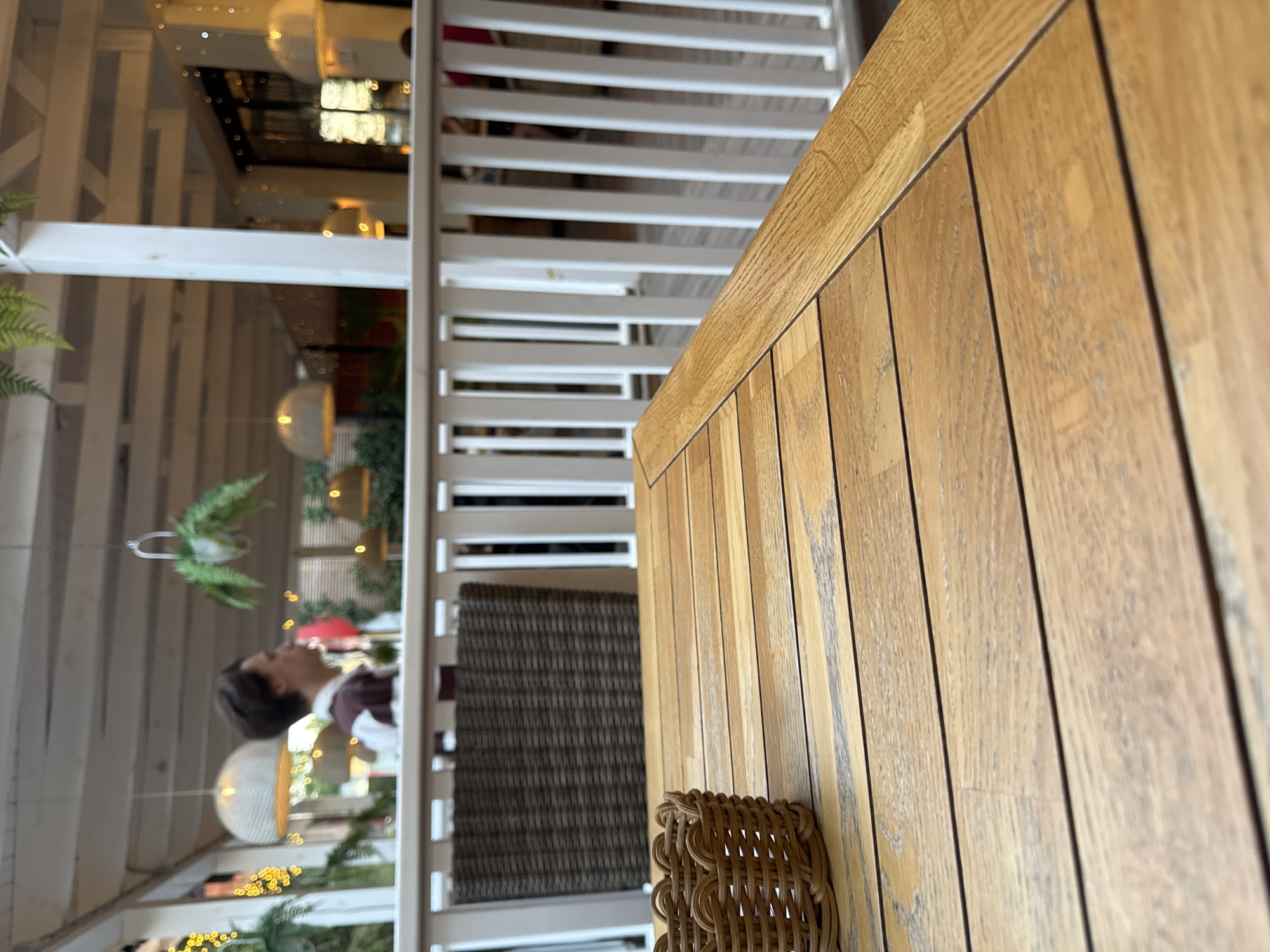 The image captures a close-up view of a wooden table in an outdoor seating area, likely a cafe or restaurant, in Yevpatoriya, Ukraine. In the background, a person with short, dark hair is visible, wearing a maroon and white striped shirt, and looking to the left. Beyond the white railing, other tables and chairs are sparsely populated. Hanging lights and greenery adorn the covered outdoor space. A small, woven basket sits on the table in the foreground. The lighting suggests it is either daytime or early evening.