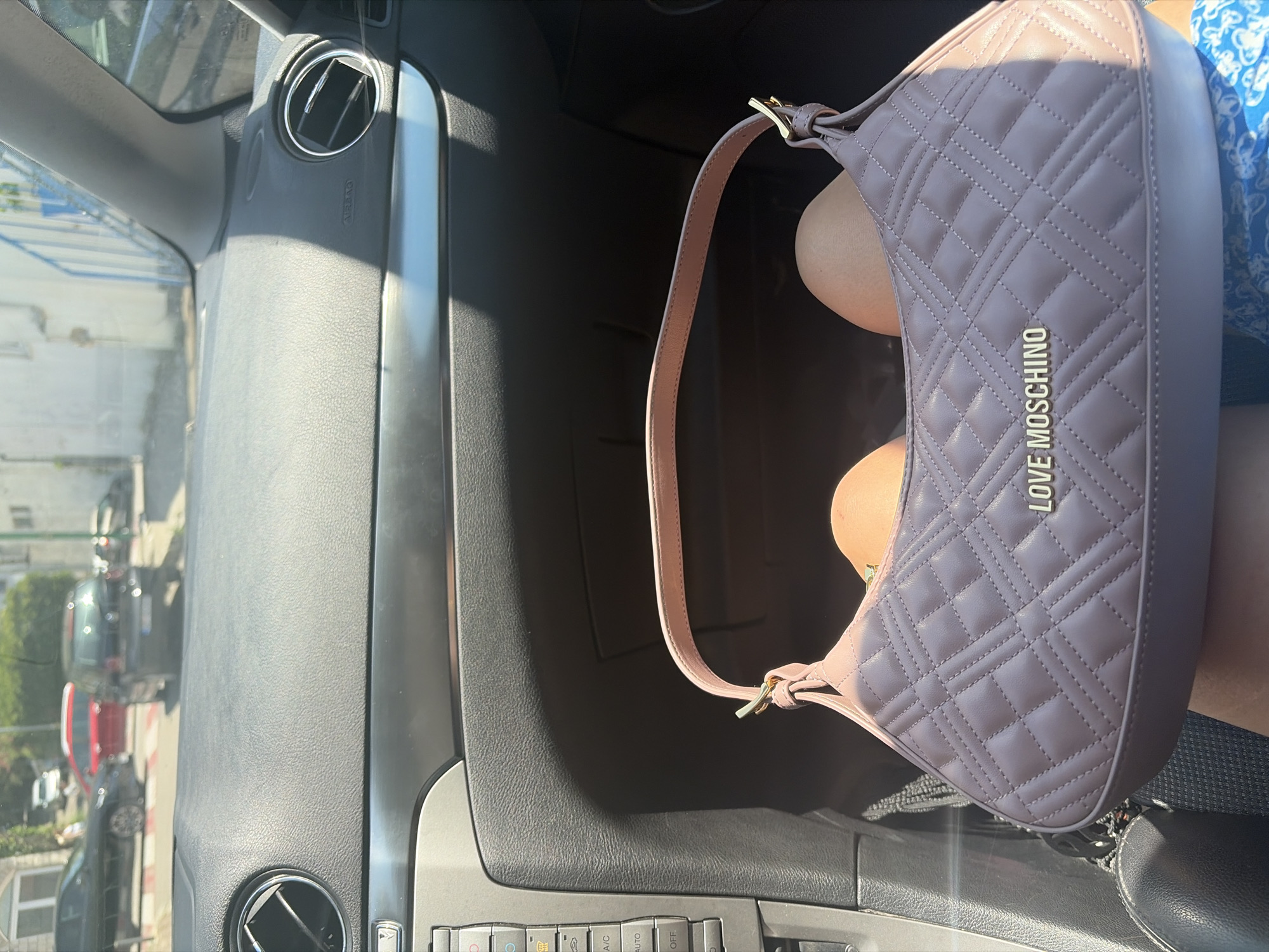 The image shows a quilted pink handbag with gold hardware and the embossed "LOVE MOSCHINO" logo, resting on a person's lap inside a car. The car's dashboard, including air vents, buttons, and a gray trim, is visible. Outside the car, through the windshield, a street scene in Budva, Montenegro is depicted, with several cars parked and in motion, buildings, and trees under a bright sky.