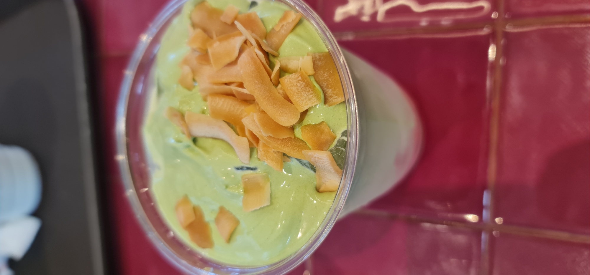 A high-angle, slightly tilted, close-up shot captures a green smoothie topped with toasted coconut flakes in a clear plastic cup. The smoothie appears creamy and thick. The coconut flakes are various sizes and shapes, ranging from small squares to longer strips.
The cup sits on a surface covered with red tiles with light-colored grout. A blurred gray object is partially visible in the lower left corner of the frame. The shot seems to have been taken indoors.