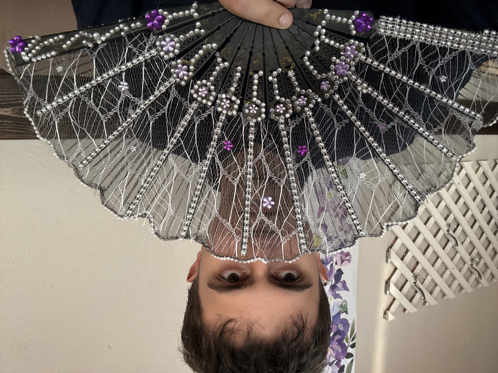 A person is holding a decorative hand fan in front of their face. The fan is black with white lace, silver bead accents, and purple flower details. The person has brown hair and brown eyes. The background includes a light-colored wall, a patterned floral design, and a white lattice structure. There is no information present to indicate this image was taken in Petarch, Bulgaria.