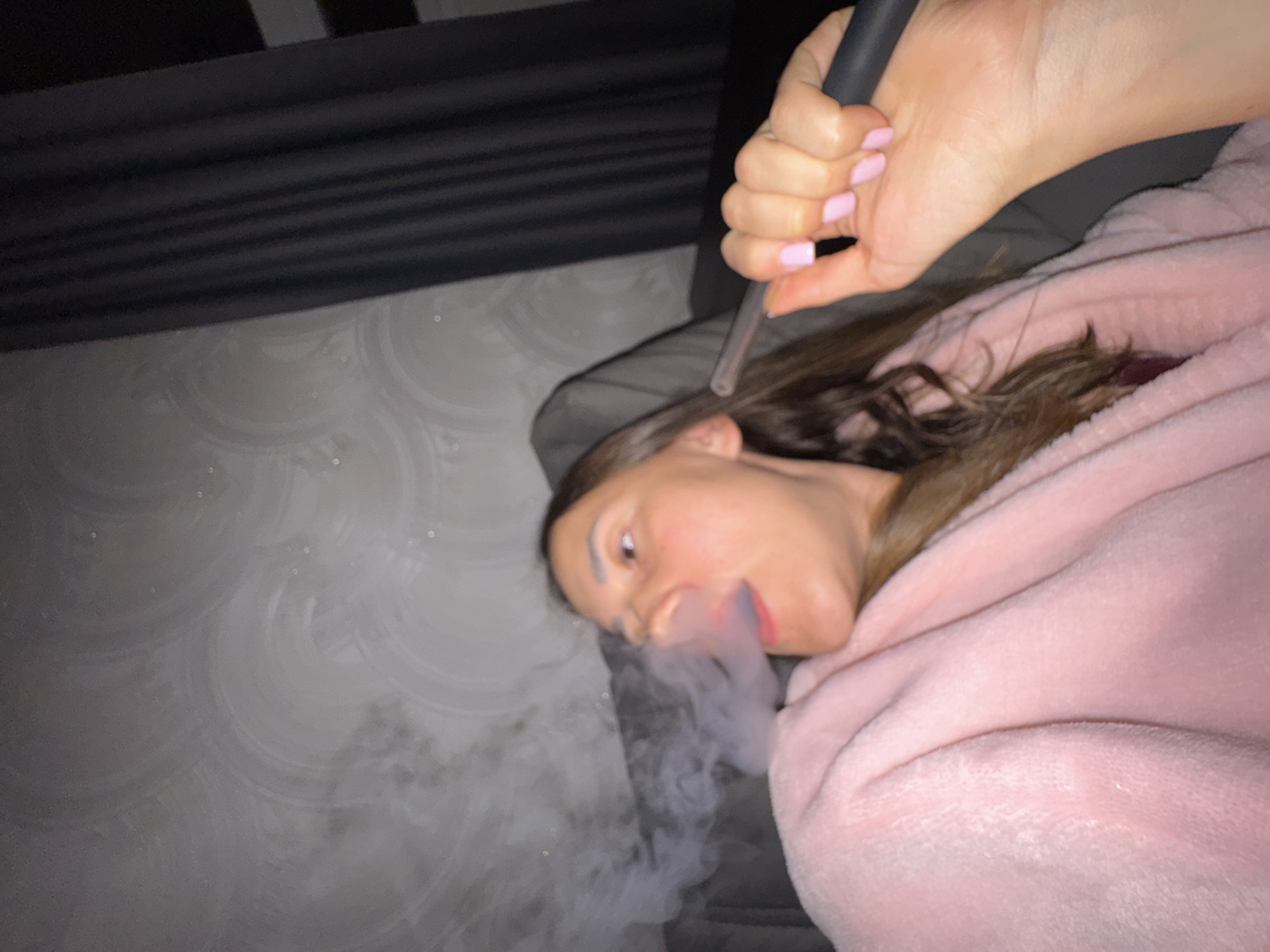 A young adult female is depicted in a dimly lit indoor setting, reclining on what appears to be a dark gray or black sofa or armchair. She has brown hair and is wearing a pink, plush robe or blanket. Her right hand, with nails painted pink and a black tip on the index finger, holds a clear and dark-colored mouthpiece of what appears to be a vaping device or small hookah. She is exhaling a visible cloud of white vapor from her open mouth, looking towards her right (viewer's left). The background features a light-colored wall with a subtle, circular or swirling pattern wallpaper, possibly white or light gray, with small reflective specks. Dark gray curtains are visible on the right side of the image. There are no identifiable features within the image that confirm the specific location as Sochi, Russia.