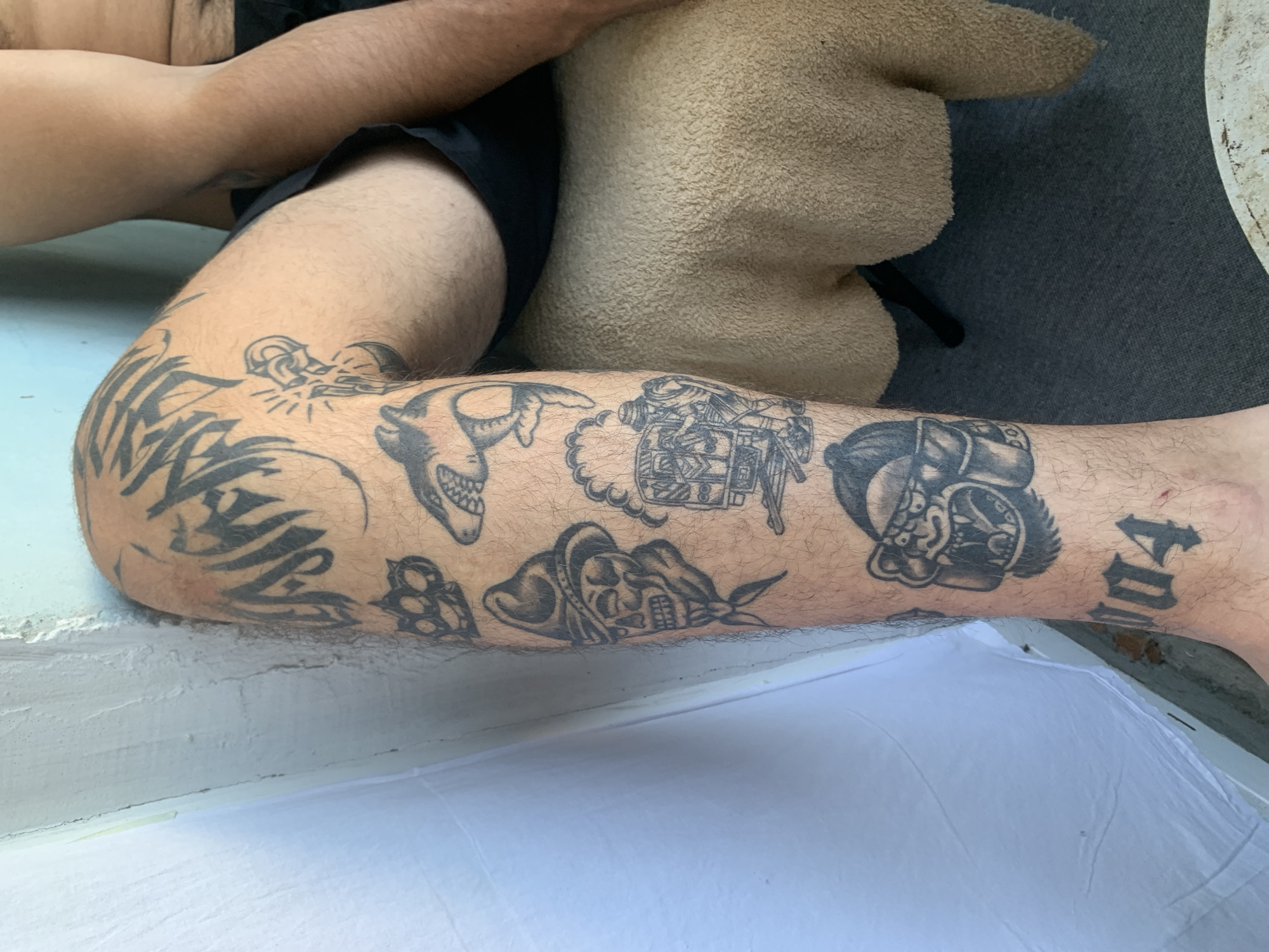 The image shows a person's legs with several tattoos. The tattoos include a shark, a skull wearing a bandana, a pair of brass knuckles, a mechanical or robotic head, and some stylized lettering that may say "104". The person is wearing black shorts. The background suggests an outdoor setting, possibly a patio or a poolside area, with a textured beige object that looks like a stuffed animal or cushion. There is also a white surface visible at the bottom of the frame, which could be a piece of fabric or furniture. The lighting indicates it is daytime.