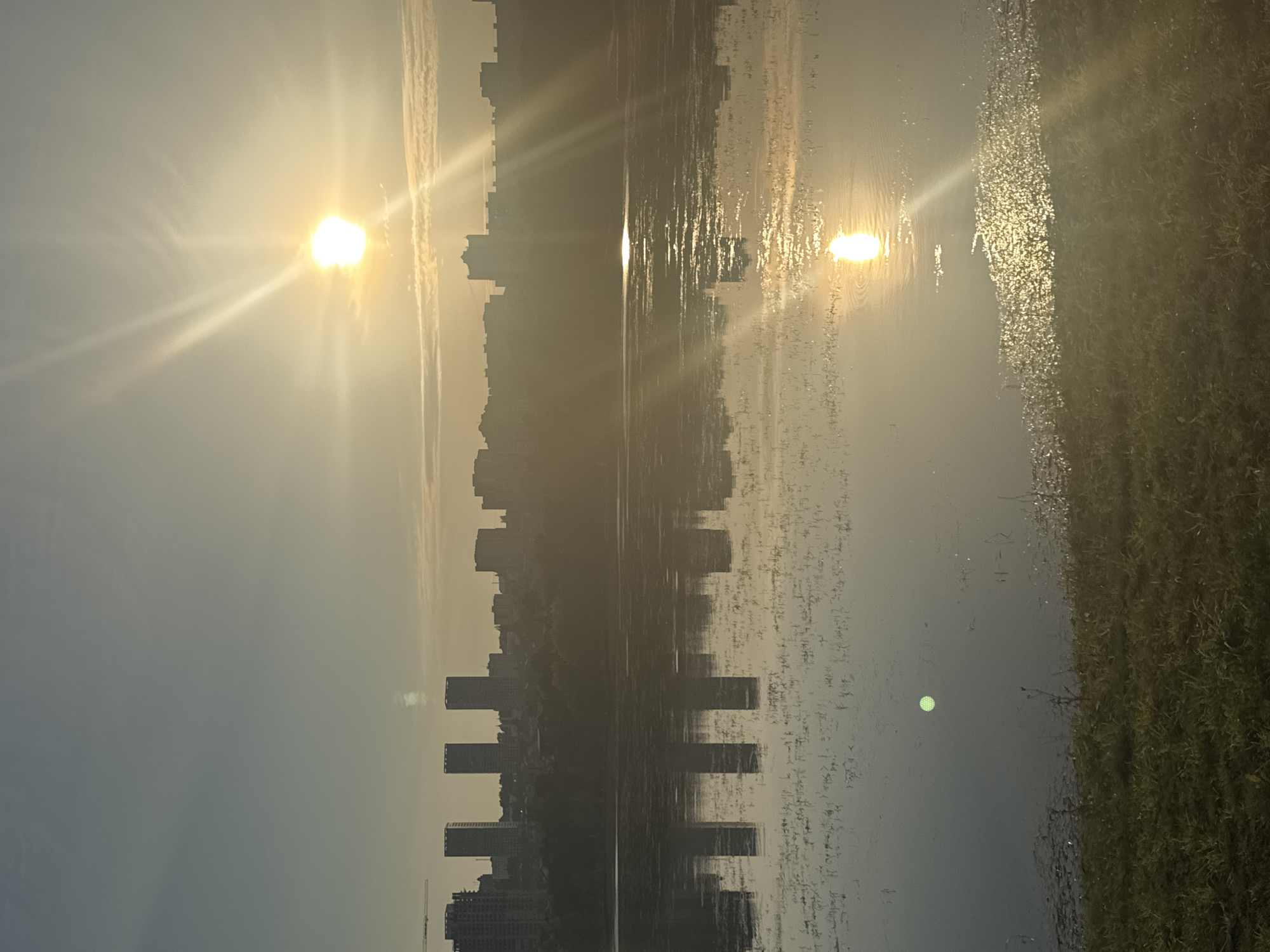 The image depicts a panoramic view of the city of Ufa, Russia, at sunrise. A bright sun is visible in the upper right portion of the sky, casting a warm glow and creating light rays that extend across the scene. The sun's reflection shimmers on the surface of a body of water, likely the Belaya River, which dominates the foreground. The water's surface is partially covered with sparse vegetation. In the middle ground, a silhouetted cityscape stretches across the horizon, featuring numerous buildings, including several tall, rectangular structures that appear to be apartment buildings or office towers. Trees and other greenery are visible between the buildings and the water. The sky above the cityscape is a pale blue, with a few wispy clouds. The overall impression is one of a serene and developing urban landscape bathed in the soft light of early morning.