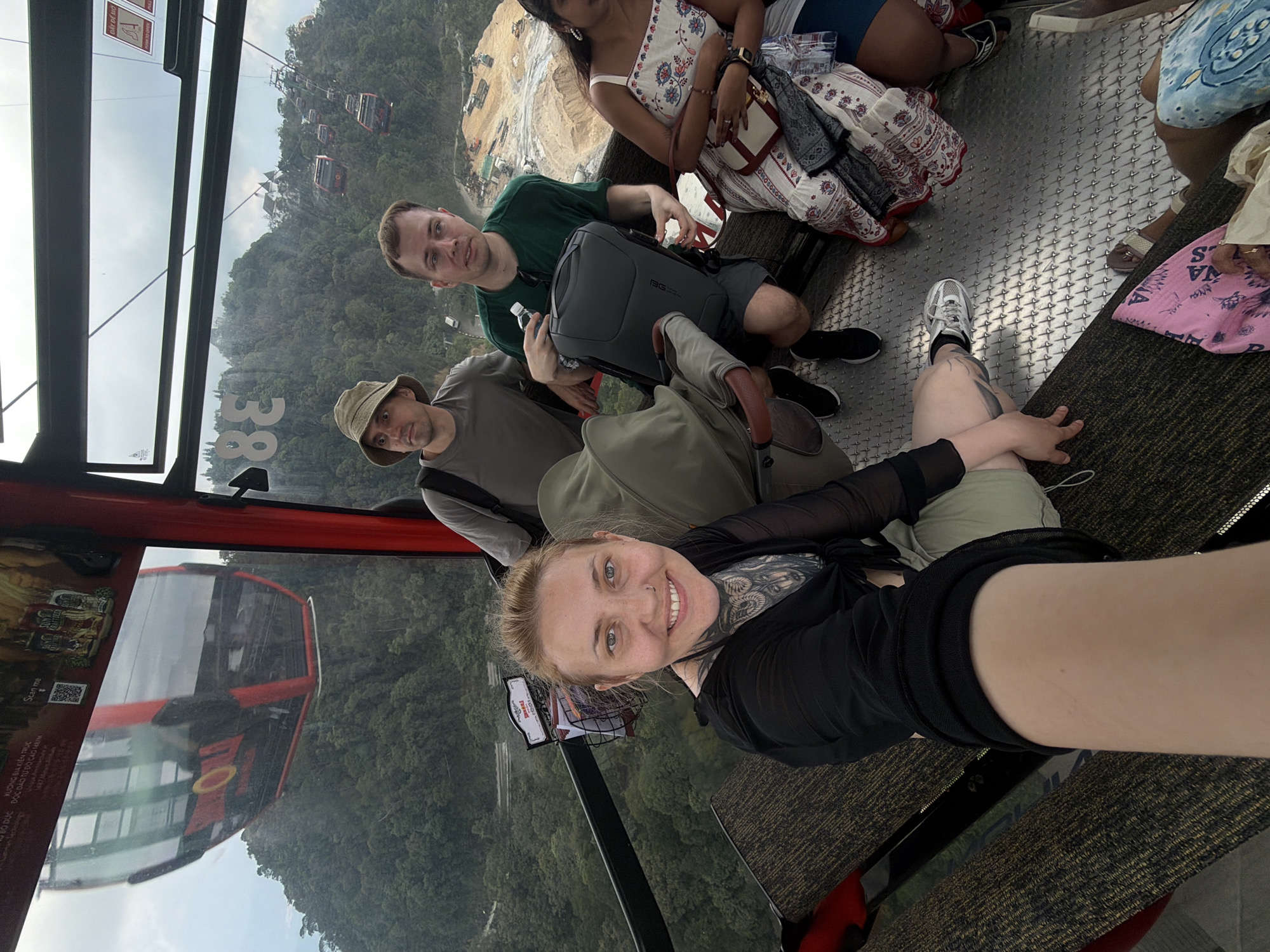 The image captures the interior of a cable car ascending a mountainous region, likely in Đà Nẵng, Vietnam, as evidenced by the Vietnamese language on a promotional poster within the car. There are at least five people in the cable car. In the foreground, a woman is taking a selfie. She is wearing a black top and light green pants. She has tattoos on her neck and chest and is smiling at the camera. Behind her are a man in a green shirt, a man in a gray shirt and khaki hat, and two women wearing dresses. A gray backpack sits between the two men. In the background, the surrounding landscape is visible through the car windows. Dense green trees cover the mountain and the cables and cars of the cable car system are visible outside the car.