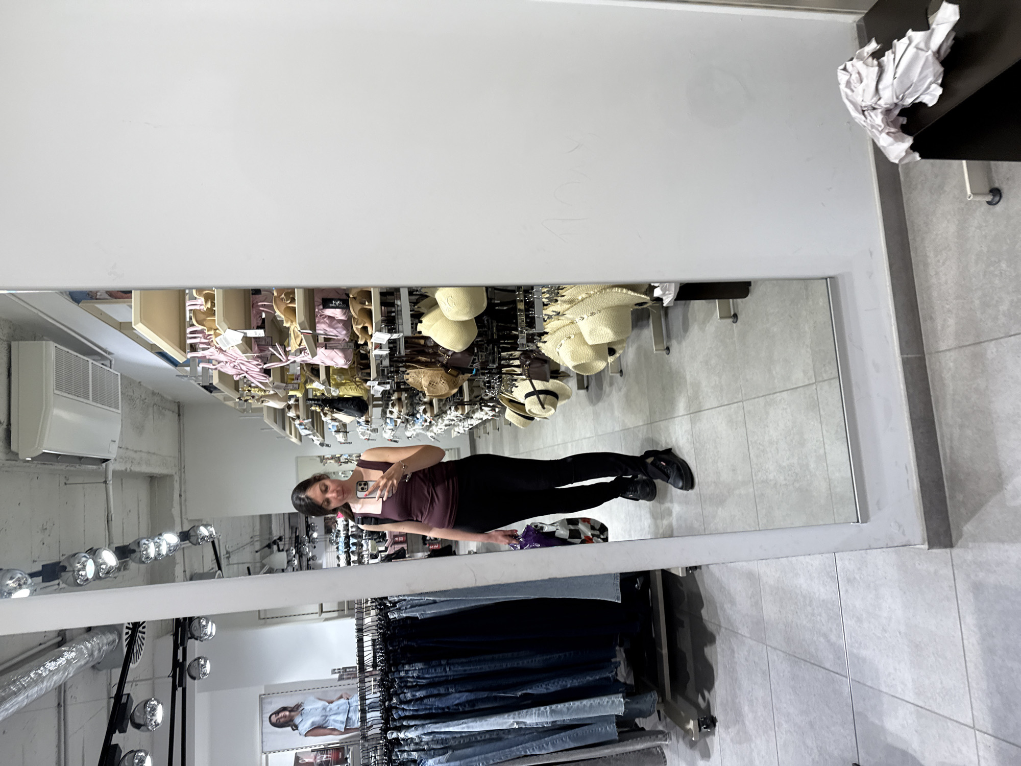 In a retail store, a woman stands in front of a full-length mirror, taking a selfie with her phone. She is wearing a dark maroon tank top and black leggings, with chunky black shoes. She is holding a multi-colored shopping bag in her left hand. The mirror reflects rows of clothing on racks to her left, and shelves stocked with accessories, including hats and handbags, to her right. The store's ceiling features track lighting and air conditioning vents. The floor is tiled in a light gray pattern. In the background, a poster of a woman in a light blue denim outfit is visible on the left. The right side of the image is dominated by a plain white wall, with a dark table in the foreground on which a crumpled piece of white paper rests. The overall environment suggests a clothing or department store.