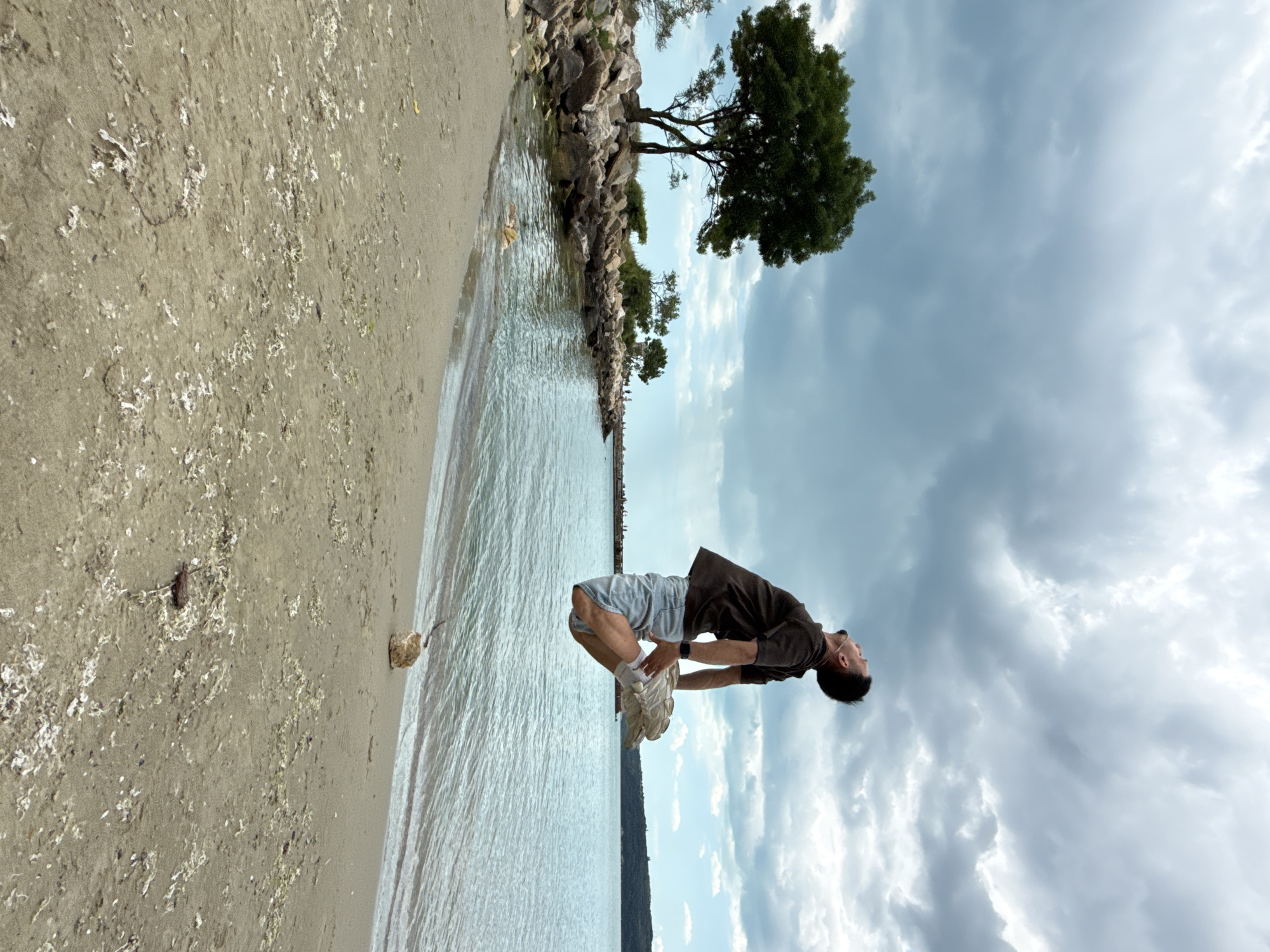 A rotated, medium shot captures a man performing a backflip on a beach. He's wearing a dark brown t-shirt, light-colored shorts, white socks, and chunky white sneakers. His arms are bent, palms facing the sky, as he arches backward. The scene is framed by the shoreline on the left, with a sandy beach transitioning into a body of water, and the sky filled with clouds on the right. In the background, along the shoreline, is a rock barrier with some trees. This is most likely the northern shores of the city of Varna, Bulgaria.