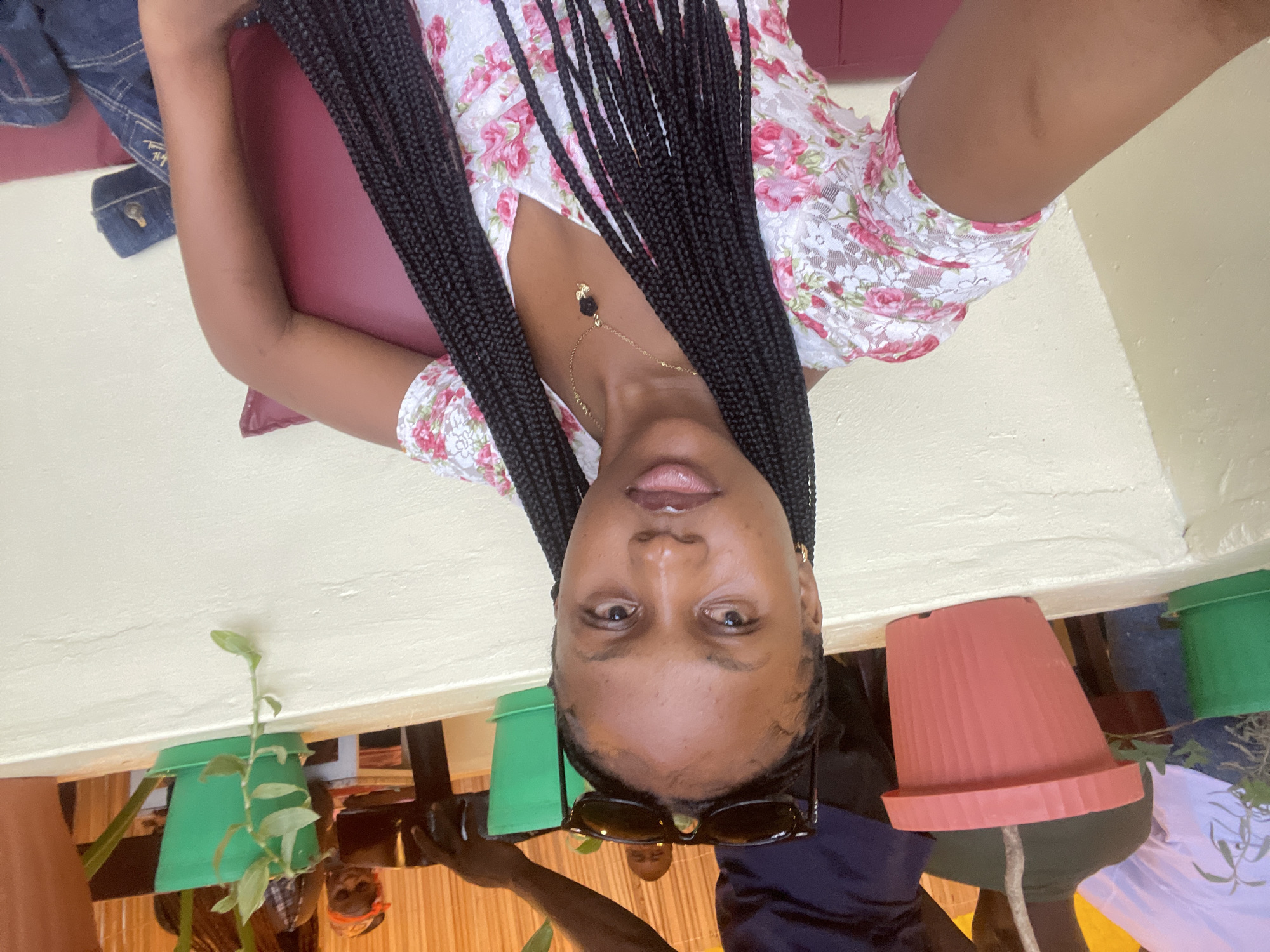 The image is a medium shot selfie of a young Black woman with long, braided hair, wearing a white floral print dress. She is seated, leaning against a burgundy cushion, and is wearing a gold necklace with a black rose pendant. Sunglasses are perched on top of her head. In the background, several potted plants are visible, including a large pink pot and a smaller green pot. Behind the plants, there is a wall made of tan-colored, rough-textured material. In the further background, partially visible, are two adults and a child. The woman's posture and the presence of a pillow suggest she is in a relaxed, outdoor or semi-outdoor setting, possibly a patio or garden area in Gura Top, Nigeria.