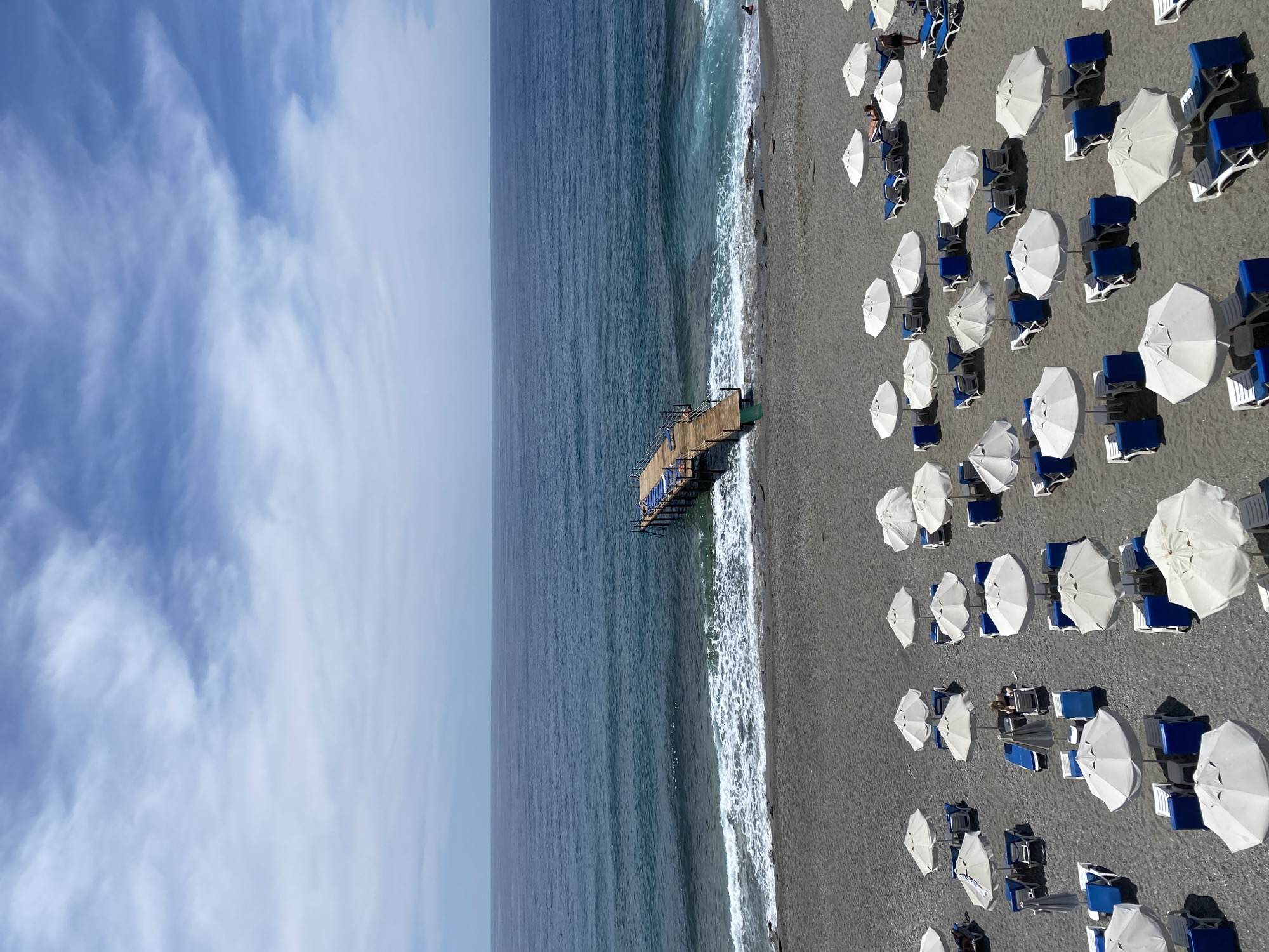 The image captures a bird's-eye view of a beach, likely in Alanya, Turkey, given the distinctive features of the landscape. A pier extends from the shoreline into the blue sea. The beach is populated with closely arranged white umbrellas and blue beach chairs, suggesting a resort or popular tourist spot. The sky shows scattered clouds. The water is relatively calm.