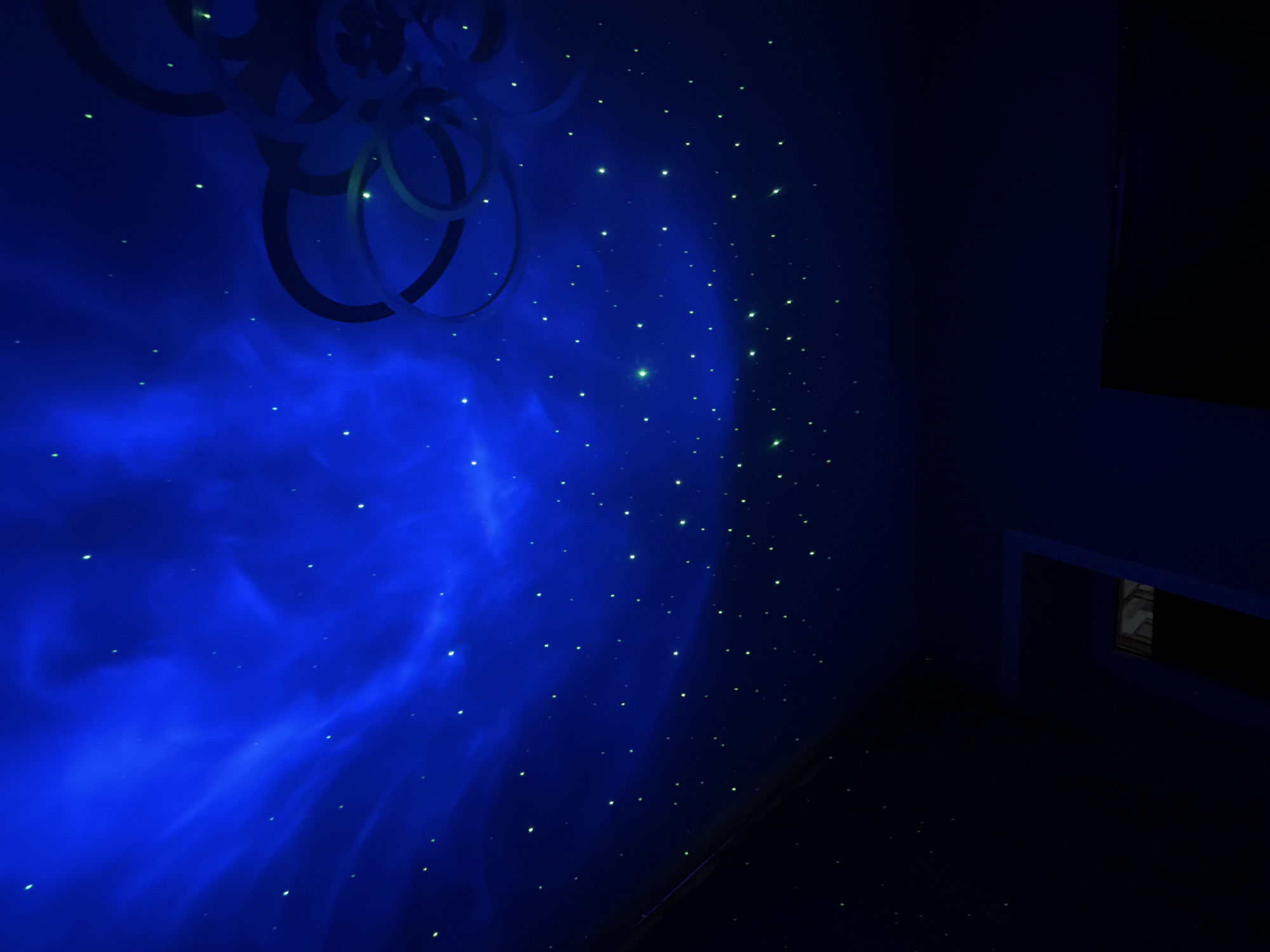 The image shows a room with a dark blue aesthetic, possibly a bedroom or entertainment room. The left wall is illuminated with a swirling, nebulous blue light, along with small, evenly spaced green lights resembling stars. A decorative circular design is mounted on the same wall. The room is dimly lit, with a piece of furniture visible on the right side of the frame. The presence of star-like lights and the nebular projection suggests the use of a star projector. The photograph doesn't provide information about the location in Reshetnikovo, Russia, or about people, actions, or interactions within the scene.