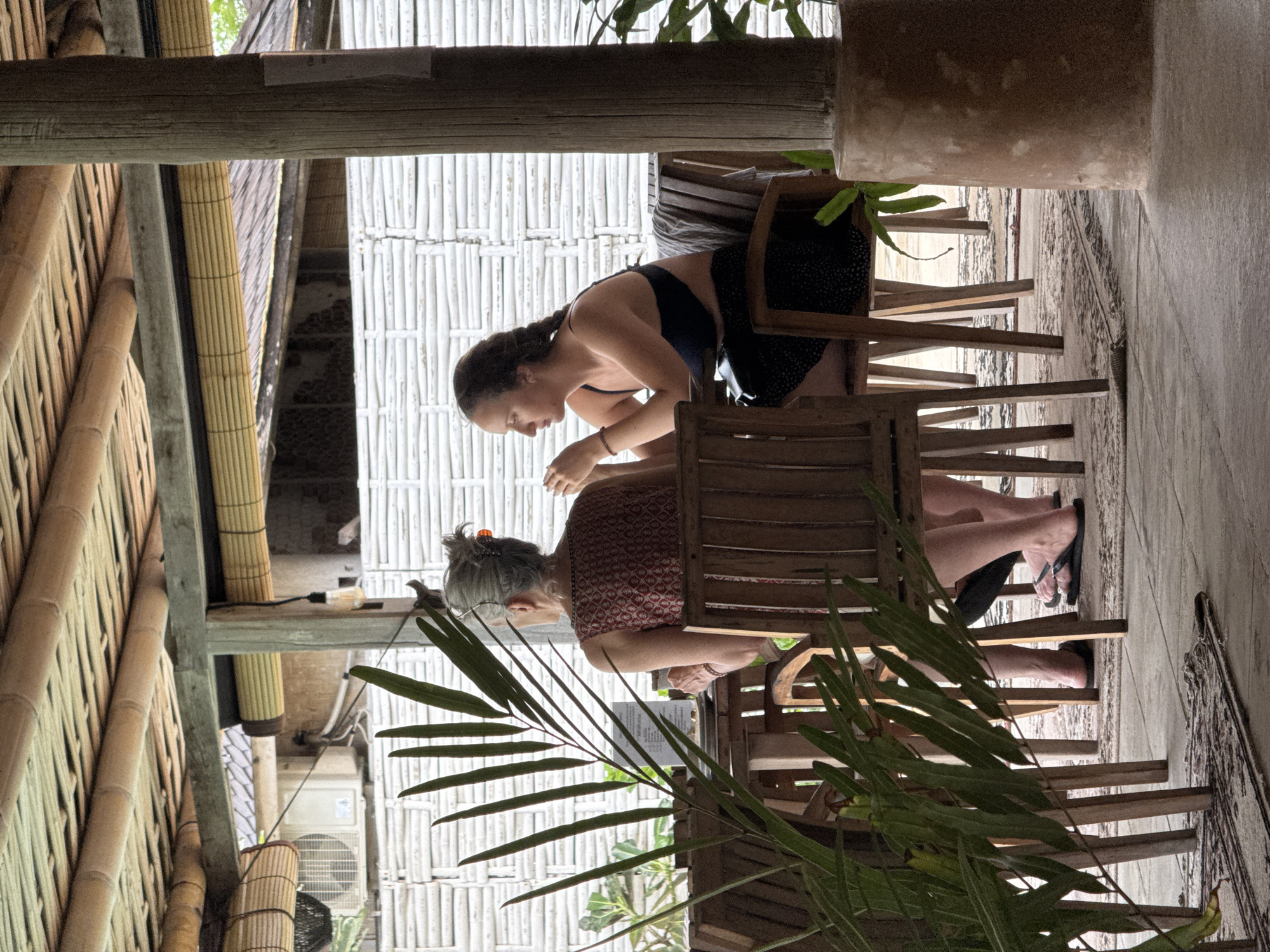 Two women are seated at a wooden table, possibly in a restaurant or cafe. One woman is younger, wearing a dark top and a black skirt with white polka dots. She is leaning forward, looking at something on the table. The other woman is older with short grey hair and is wearing a patterned red and white top. She is also looking down at the table. The table is made of wood with thin slats and there is a palm frond in the foreground obscuring part of the view. The structure behind them appears to be constructed from bamboo and other natural materials, suggesting a tropical location. There is an air conditioning unit visible in the background. A building can be seen in the distance beyond the wall. The location is Unknown, Indonesia.