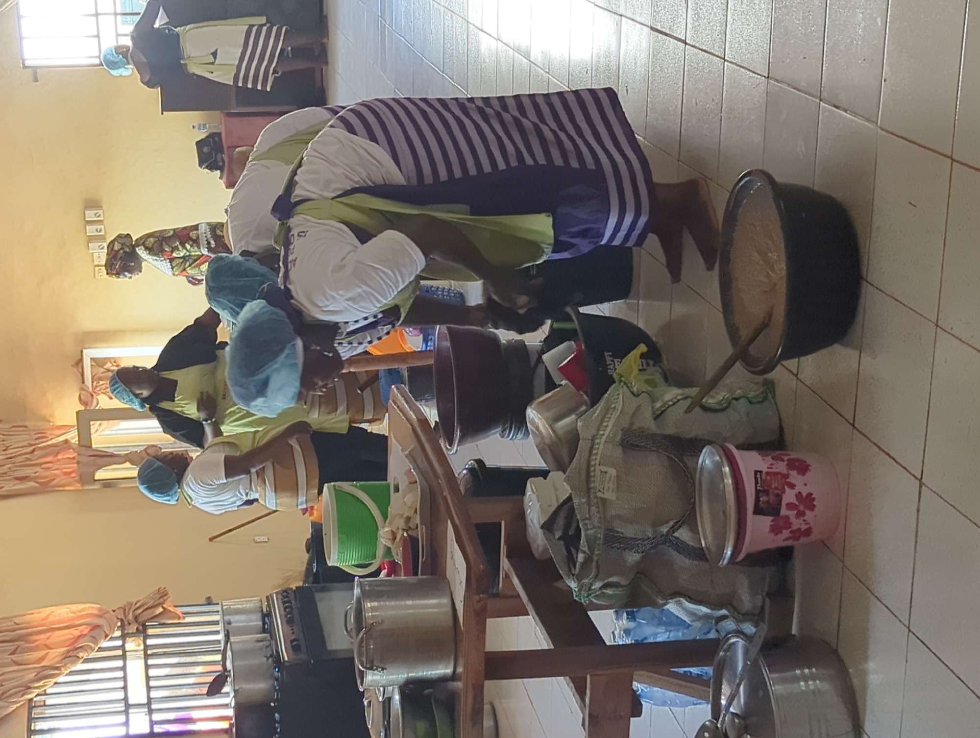 The image shows a group of people indoors, seemingly in a kitchen or food preparation area. There are at least five individuals visible, all wearing head coverings, and some have aprons. They appear to be involved in preparing food, with various containers, pots, and pans scattered around. A large pot with a substance inside is near the foreground, and a wooden shelf holds various kitchen items. The location seems to be a functional space, possibly in the city of Unknown, Unknown, given the context of food preparation and the presence of cooking equipment. The scene suggests a communal or organized effort to prepare a meal, possibly for a large group or event.