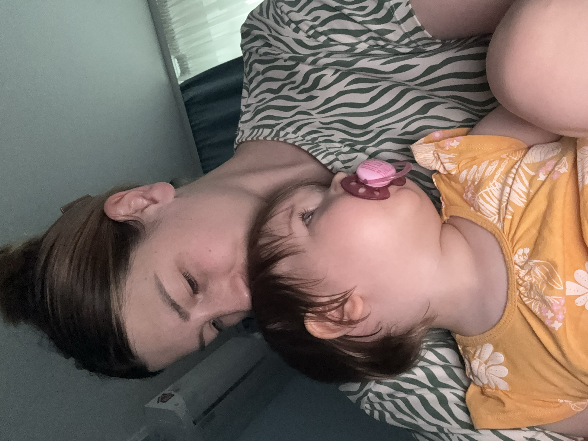A woman and a baby are lying on a bed covered with a green and white zebra-striped sheet. The woman has brown hair pulled back in a bun and is lying on her side with her eyes closed. The baby is lying on its side with a pink pacifier in its mouth and is wearing a yellow shirt with white floral designs. The location is not identifiable as Moscow, Russia from the image.