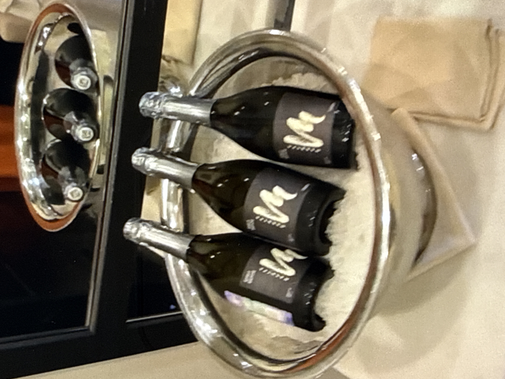 Three small dark glass bottles, each with a metallic silver foil-covered neck and a black label with a stylized "M" in a light color, are arranged in a large, shallow silver bowl filled with ice. The bowl is placed on a light-colored surface, possibly a table covered with a cloth. In the background, a shiny silver tray holds three more of the same bottles, partially obscured. A dark, reflective surface, possibly a mirror or a polished table, is visible behind the bottles. A folded light brown napkin is positioned to the right of the ice-filled bowl. The lighting is dim and warm.
