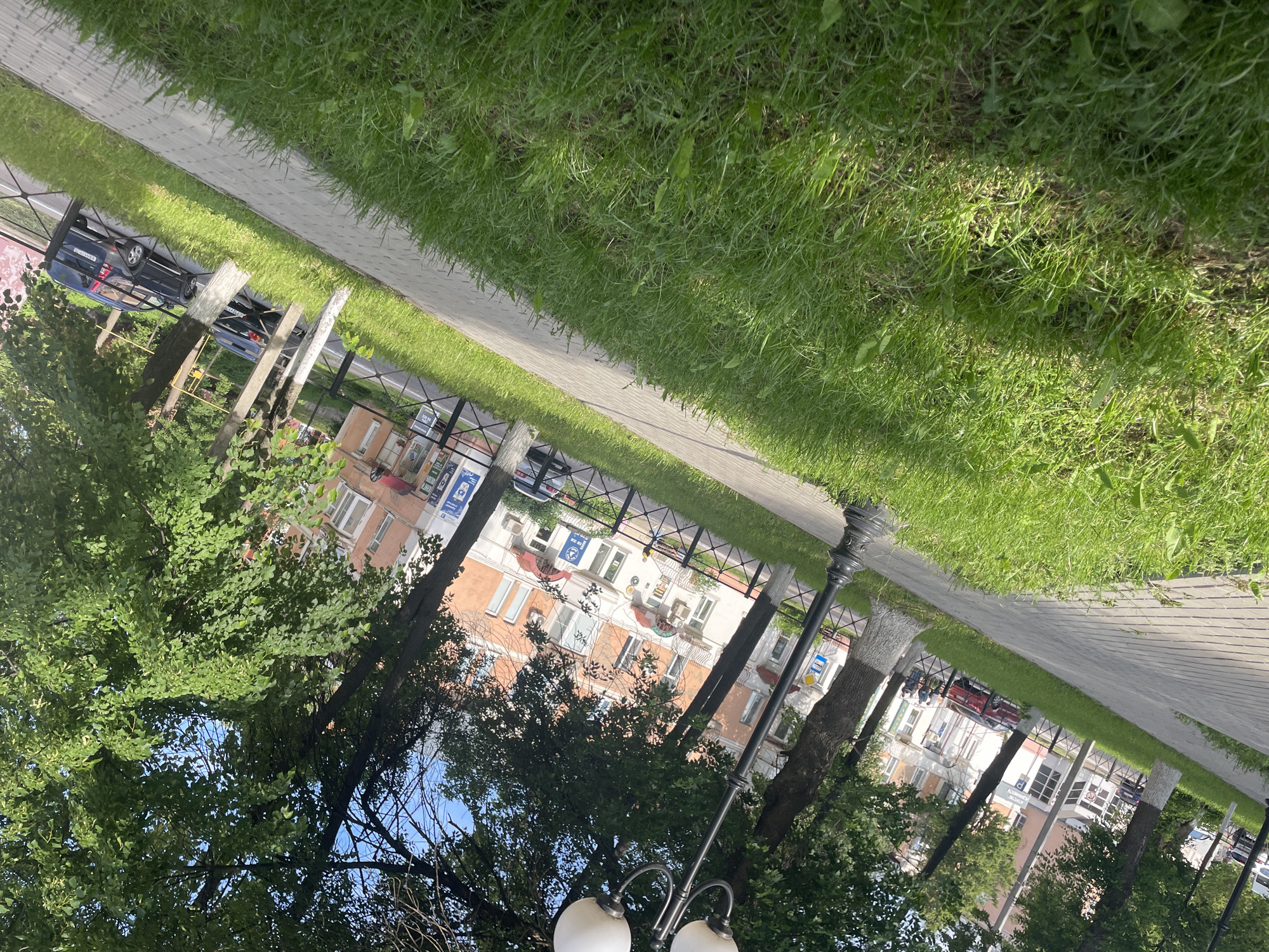 This media file, when viewed in its correct orientation, displays a sunny daytime scene from a city park or street in Votkinsk, Russia. The foreground features a grey paved sidewalk running alongside a strip of vibrant green grass.
Mature trees with abundant green foliage line the sidewalk, partially obscuring the background. Beyond these trees and a black metal fence, a street is visible with several parked cars. A dark blue sedan with a Russian license plate "A344EE" is clearly identifiable. In the background, multi-story buildings stand, characterized by multiple windows, some balconies, and numerous external air conditioning units. Several signs are affixed to these buildings, including blue ones with white Cyrillic text, partially legible as "Ремонт" (Repair), "АТЕЛЬЕ" (Atelier/Workshop), and "СЕРВИС" (Service), indicating commercial businesses.
A decorative black street lamp with two globe-shaped lights is prominent in the right foreground. The sky is clear and blue, suggesting fine weather. The scene captures a tranquil urban environment, devoid of visible people or any specific ongoing activity.