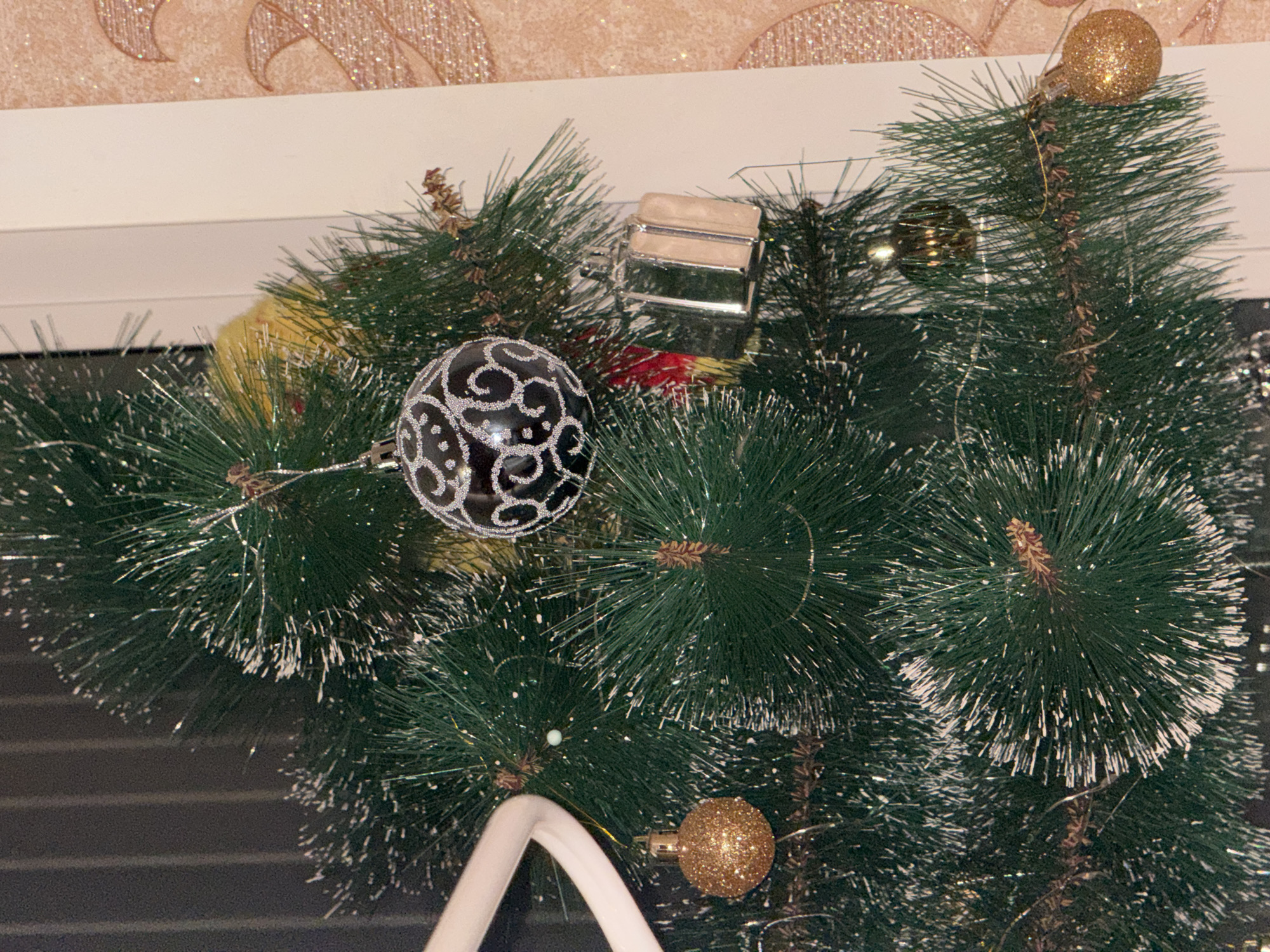 The image shows the top portion of a decorated Christmas tree. The tree is artificial, with green needles dusted with artificial snow. The decorations include a black and silver patterned bauble, a small silver box-shaped ornament, and several gold glittery baubles. A white, curved object, possibly a part of a larger decoration, is also visible. The background is a wall with a decorative pattern and a white window frame. The setting appears to be indoors. The location in Izhevsk, Russia, is not discernible from the image alone.