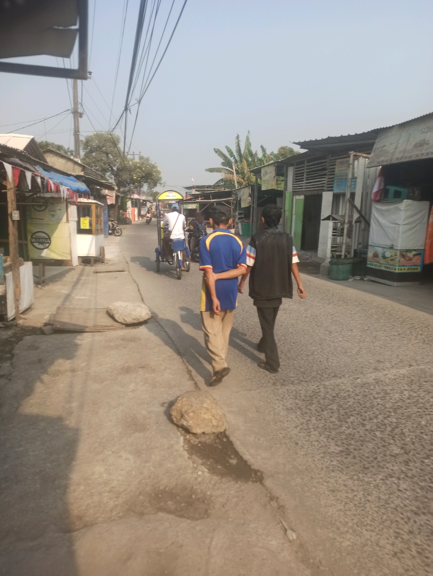 Two young males are walking away from the camera down a paved road in what appears to be a residential or commercial area in Telagamurni, Indonesia. The male in the foreground on the left is wearing a blue t-shirt with yellow and red trim on the sleeves and collar, and tan pants. The male to his right is wearing a black vest over a dark shirt and black pants. They are walking on the right side of the road.
The road is lined on both sides with buildings. On the left, there are several structures with corrugated metal roofs, some appearing to be shops or market stalls. One stall has a yellow banner with the word "WARNA" in red text. A person is riding a bicycle with a trailer in the distance on the road, and another motorcycle is parked on the left.
On the right side of the road, there are more buildings with corrugated roofs. One building has a white tarp covering a section and displays what appears to be an advertisement with text that includes "0858 5735 0852 08161-2050". There are also trees visible in the background, and a clear sky overhead. The environment appears to be an urban or semi-urban setting with signs of daily commerce and pedestrian activity. The ground in the foreground shows some wear and tear, with a large rock and a wooden plank on the paved pathway to the left of the road. Overhead, numerous electrical wires crisscross the scene.