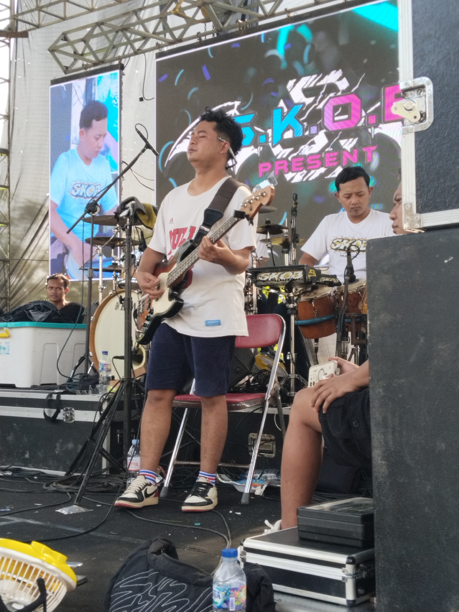 The image depicts a band performing on an outdoor stage in what appears to be Karanganom, Indonesia. The lead musician, wearing a white t-shirt, dark shorts, and sneakers, is playing a bass guitar. Behind him are two other musicians: one playing a set of drums and another partially obscured. Stage equipment, including microphones, cables, water bottles, and a small fan, are visible on the floor. A large screen displays the band's name or logo, "S.K.O. Present," and another screen shows a musician playing drums. An individual sits on a plastic container in the background observing the performance. Another individual is partly visible to the right, sitting on a black and silver case.