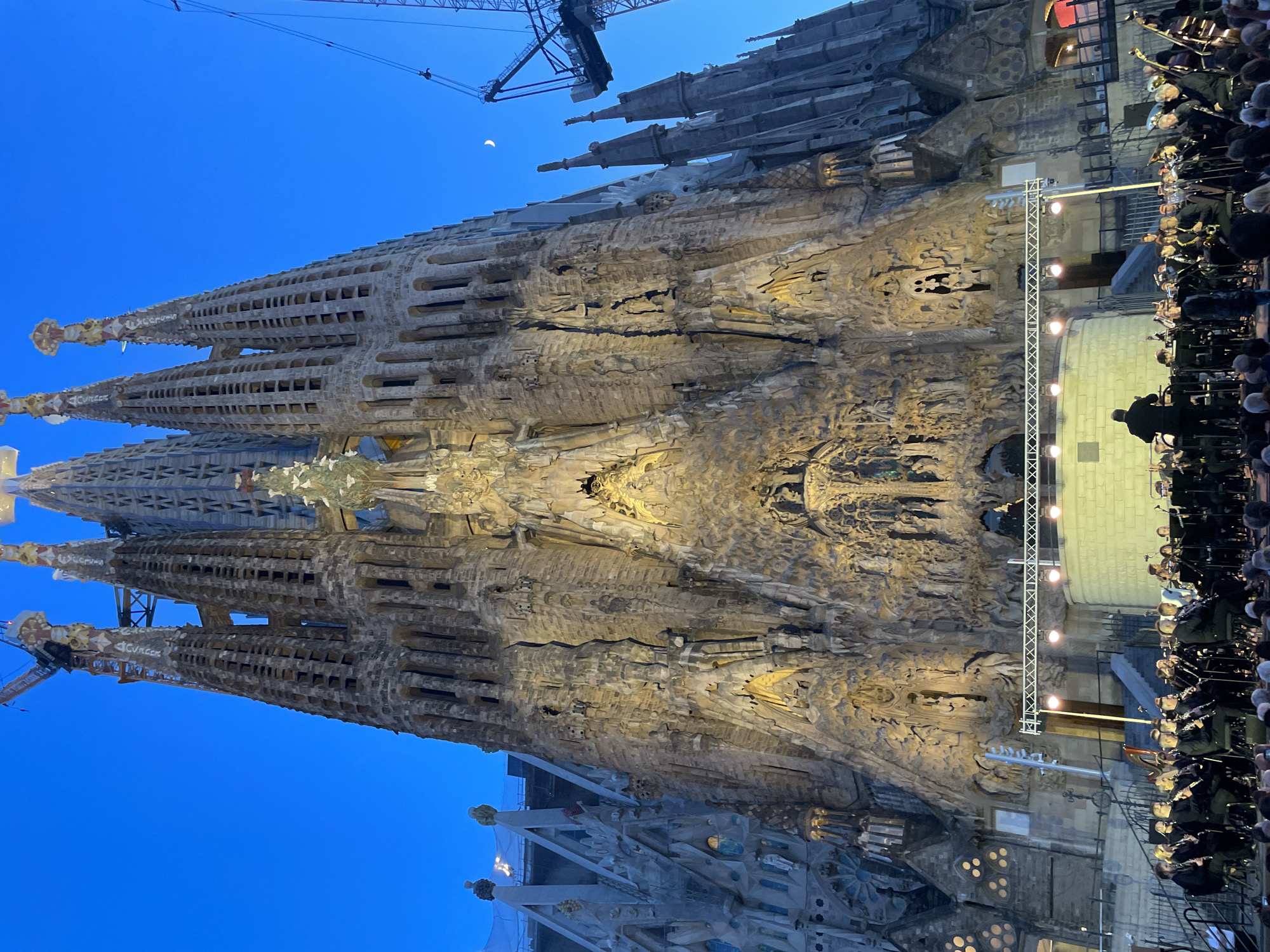The image shows a wide-angle, low-light view of the Sagrada Familia in Barcelona, Spain, at dusk. The iconic basilica's facades are illuminated, highlighting the intricate stonework and towering spires. In the foreground, an orchestra is performing on a stage set up in front of the basilica. Members of the orchestra are dressed in black attire and are playing various instruments, including brass, woodwinds, and string instruments. A conductor stands on a podium, directing the musicians. Spectators are seated in front of the stage, facing the orchestra and the basilica. A crescent moon is visible in the clear blue sky to the right of the basilica, along with a construction crane, suggesting ongoing work on the landmark. The overall scene depicts a cultural event taking place at a significant architectural site.