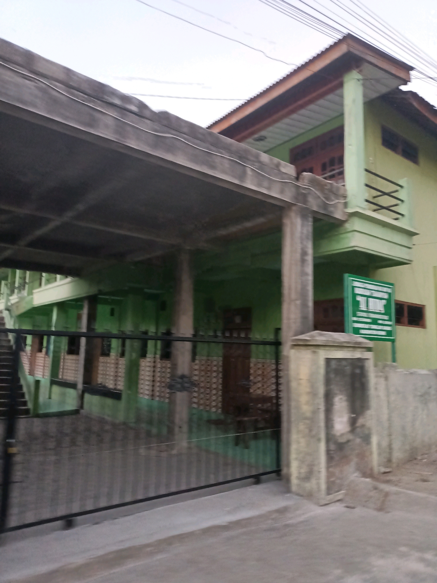 This image captures a street view of a two-story building in the city of Badas, Indonesia. The building is painted in a light green hue and features a brown corrugated metal roof. A prominent concrete overhang extends from the left side of the building, supported by columns. In front of the building, a black metal gate with vertical bars encloses a walkway. A staircase with dark treads and a white railing is visible to the left, leading up to the building's entrance. A green sign with white lettering is mounted on a post to the right of the gate, near the corner of the building. The area appears to be an outdoor space, with a concrete pathway in the foreground and some vegetation or debris visible on the right side. The sky above is overcast.