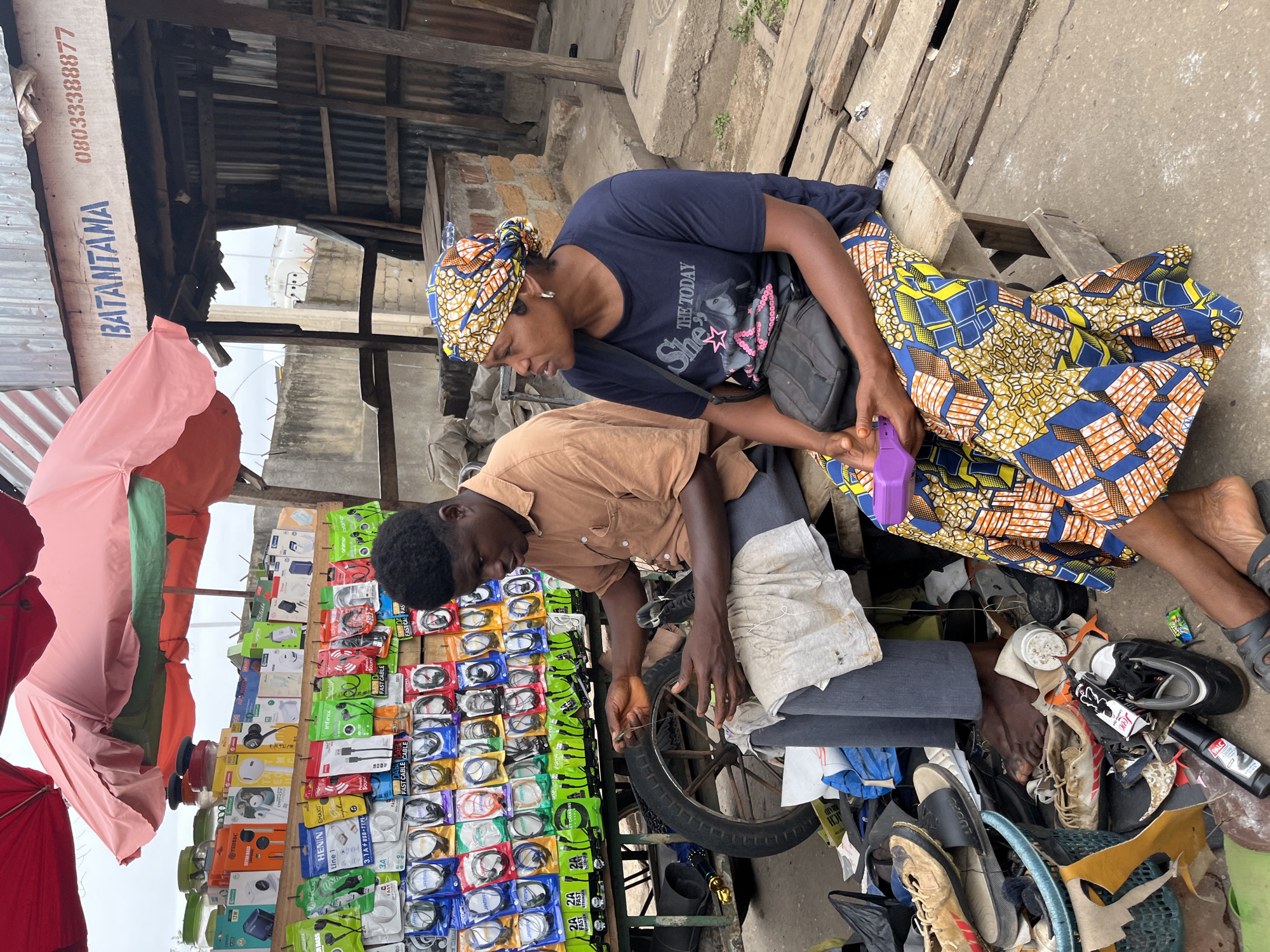 This is a full-frame, eye-level, outdoor photograph of a street vendor stall in Jos, Nigeria. The stall, covered by a pink and green umbrella, displays a variety of mobile phone accessories, including charging cables and earphones. Two people, a man and a woman, are seated on a wooden bench in front of the stall. The man, wearing a brown shirt and grey pants, is focused on repairing a shoe. The woman, wearing a dark blue t-shirt with graphics and a colorful patterned skirt, is looking to her right and holding a purple object in her hands. The ground around them is littered with shoes, a basket, and various other items. The background shows a simple building structure with corrugated metal siding and exposed wooden beams, suggesting a makeshift or informal commercial setting. The overall scene depicts a typical street market environment in Jos, Nigeria.