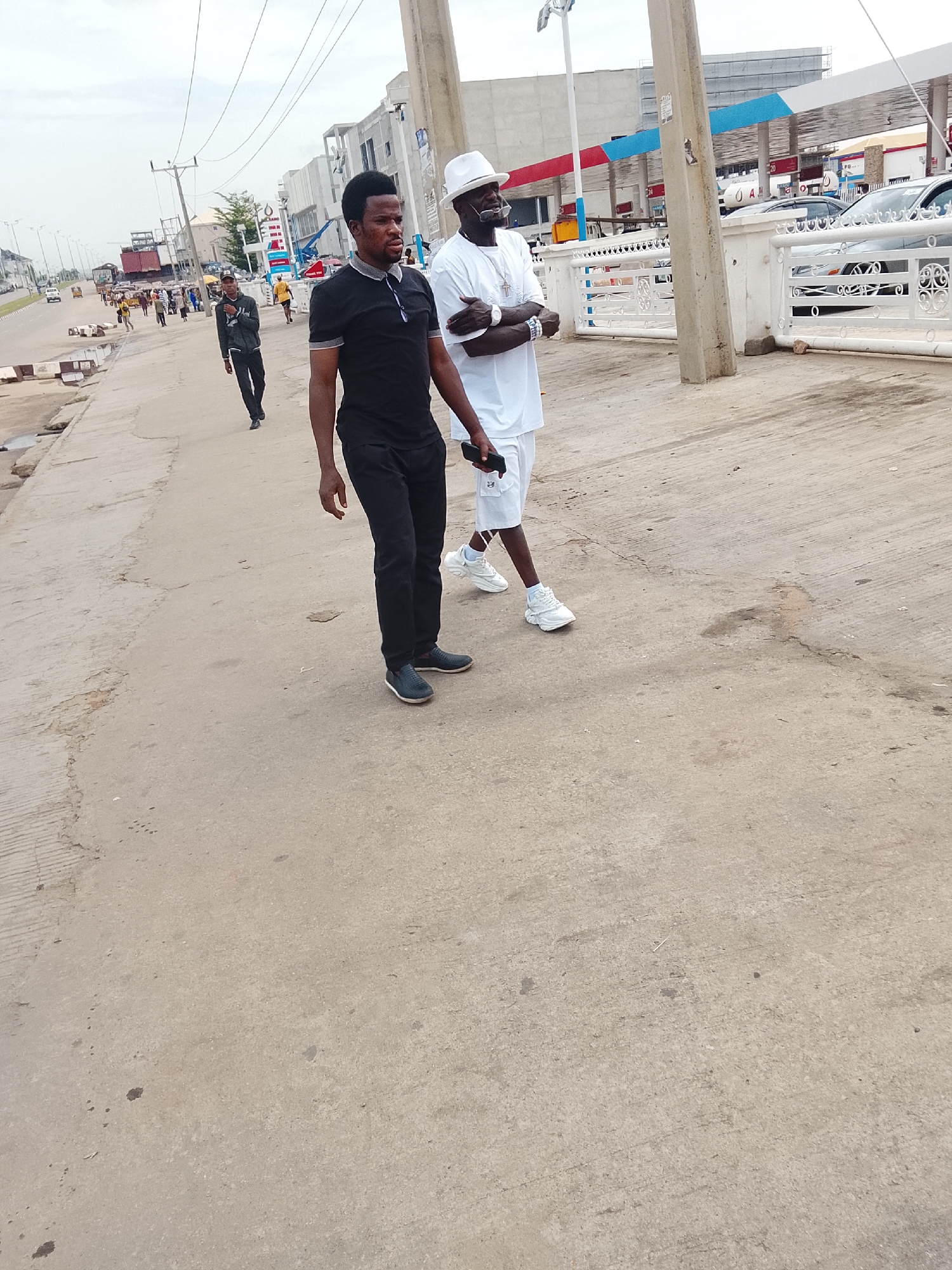 The image shows two men walking on a concrete sidewalk in what appears to be Abuja, Nigeria. The man on the left wears a black polo shirt, black pants, and dark shoes, holding a phone in his right hand. The man on the right is dressed entirely in white, including a white hat, shirt, shorts, and sneakers. He has dark sunglasses and jewelry.
Behind them is a road with sparse traffic and pedestrians. In the background, there's a gas station with red and blue accents. Overhead electrical wires span across the scene. The sky is overcast. Other people are walking along the road in the distance.