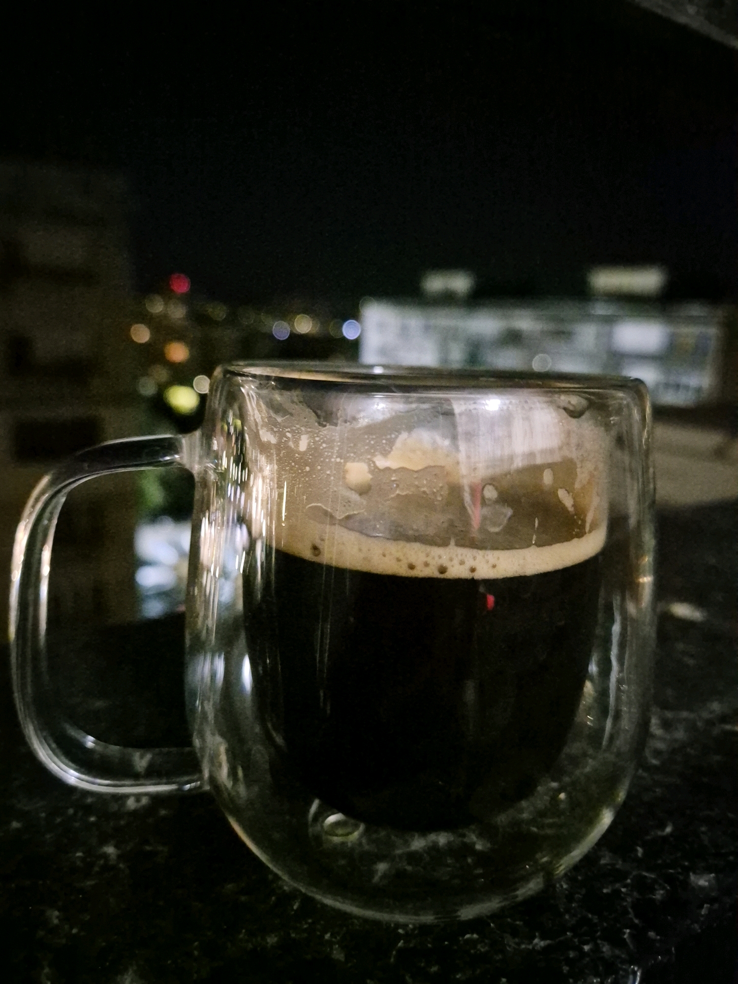 A close-up, eye-level shot shows a clear, double-walled glass mug filled with a dark beverage, likely coffee or stout, featuring a frothy head. The mug has a curved handle and rests on a dark, speckled surface. In the blurred background, city lights twinkle at night, suggesting an urban setting. The ambient light is dim, with the mug's contents and reflections being the primary sources of illumination. There are no people or discernible interactions visible in the image. The scene appears to be outdoors on a balcony or windowsill overlooking a city.