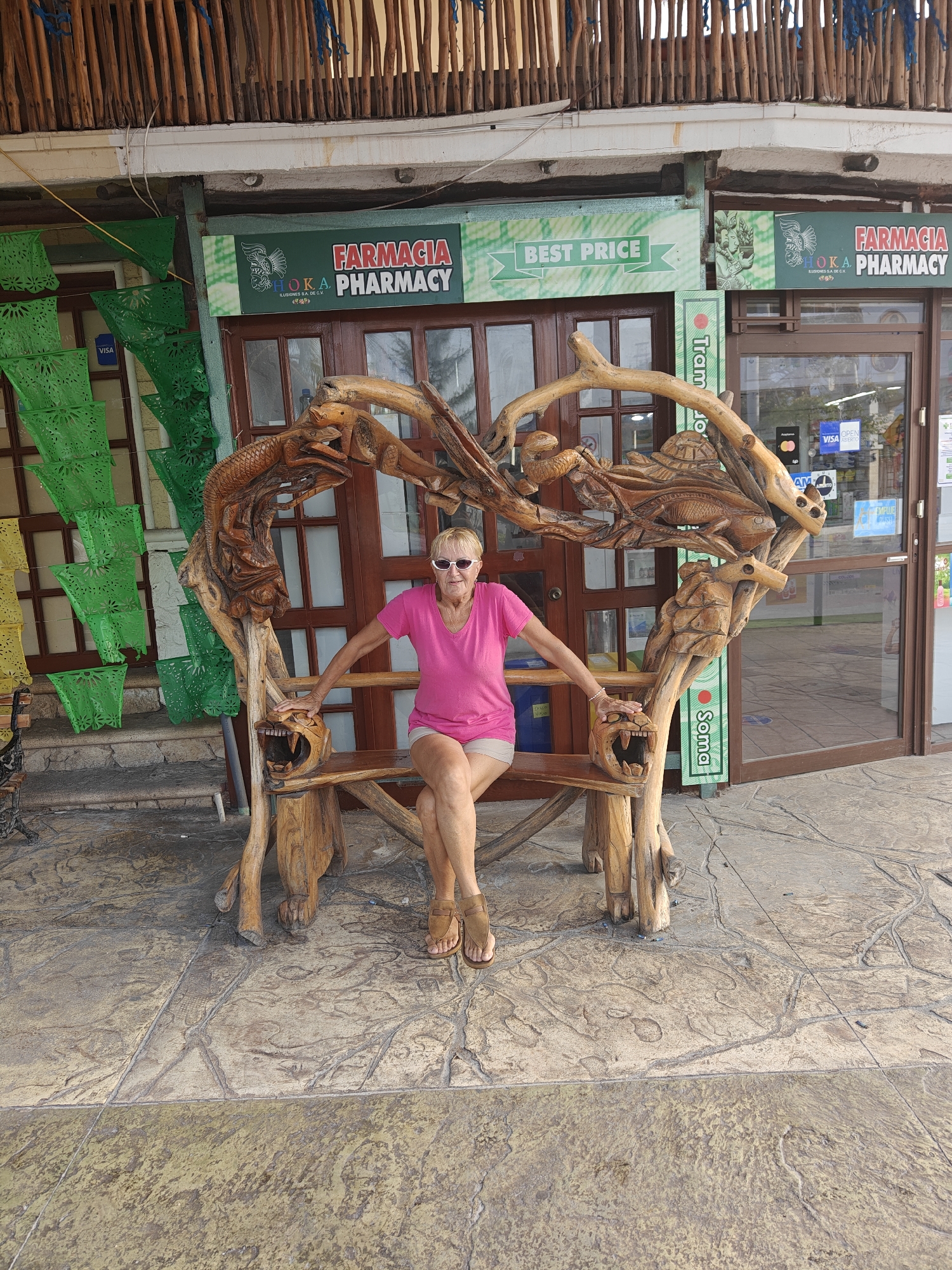 A Caucasian woman in her late 60s, wearing sunglasses, a pink t-shirt, and tan shorts, sits on a carved wooden bench outside a building. The bench is elaborately carved with what appears to be marine life and animal heads. The woman is posing with her hands on the arms of the bench, her legs crossed. In the background, there is a storefront with signs for "FARMACIA" and "BEST PRICE." The ground is paved with stone. This scene is likely in Puerto Morelos, Mexico, given the prevalence of Pharmacies and the tropical environment suggested by the decorations.
