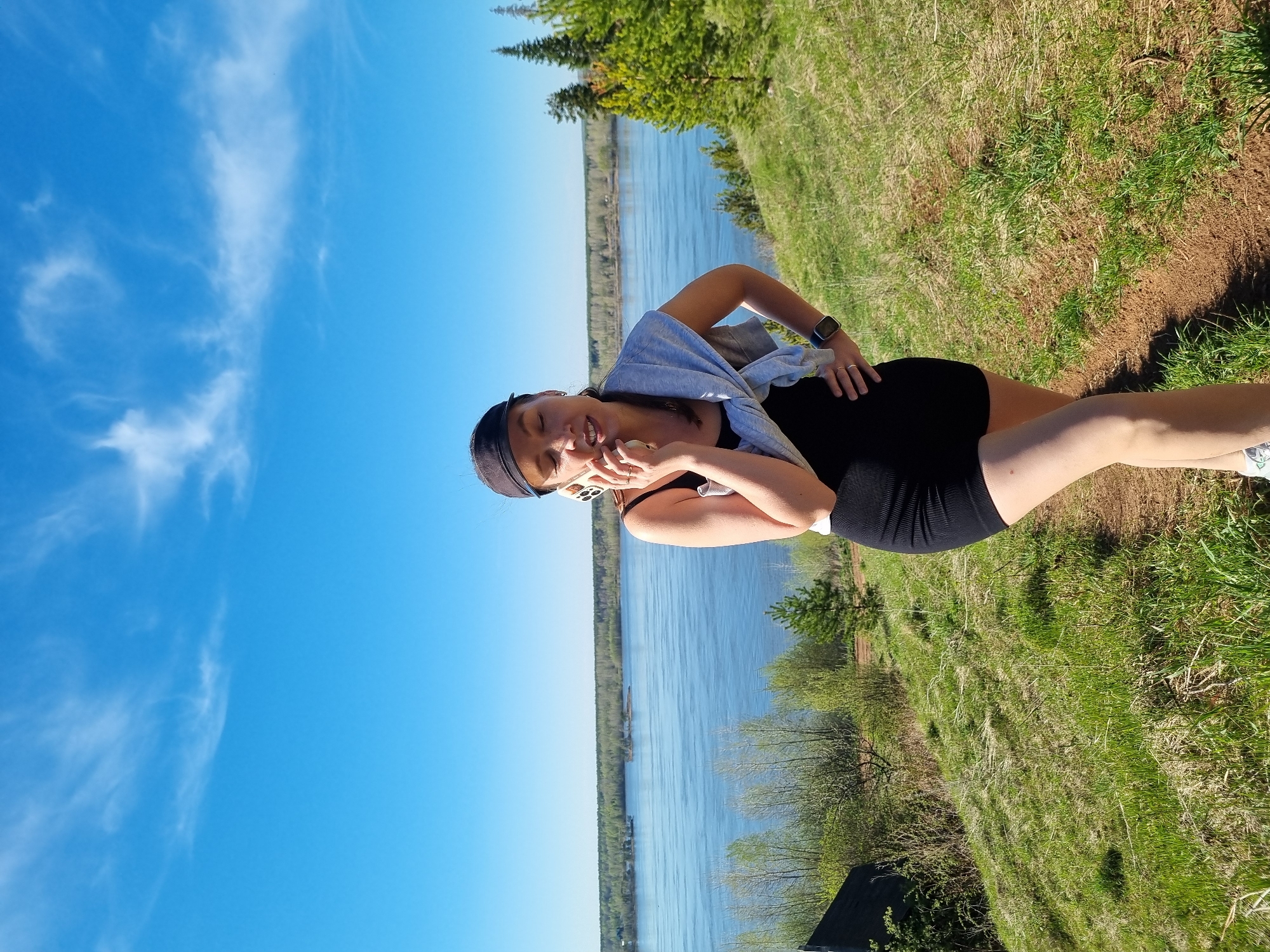 The image shows a woman outdoors near a body of water. She is wearing a black top and shorts, a light grey shirt draped over her shoulders, and a visor. She is holding a cell phone and appears to be speaking. The background includes a blue sky with wispy clouds, a river or lake, trees, and grassy terrain. The location is Ольховка, Russia.