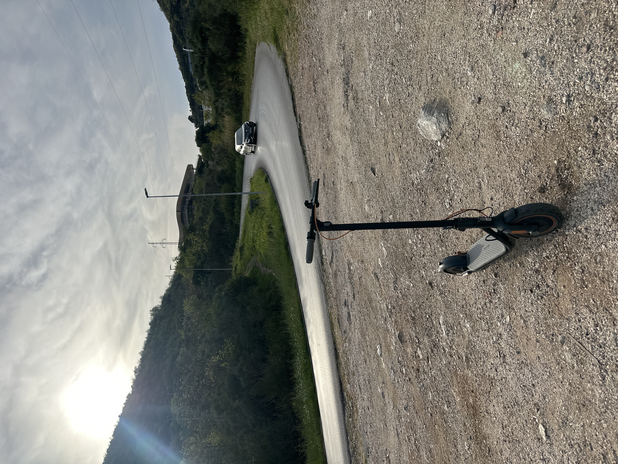 The image depicts an electric scooter lying on a gravelly shoulder next to a paved road. The road curves sharply in the background, with a car visible on the curve. The surrounding environment includes a hillside covered in vegetation and utility poles with power lines. The sun is behind the mountain. The location is likely in the Budva area of Montenegro, given the terrain and infrastructure. There are no people visible in the image. The primary object is the scooter, lying on its side.