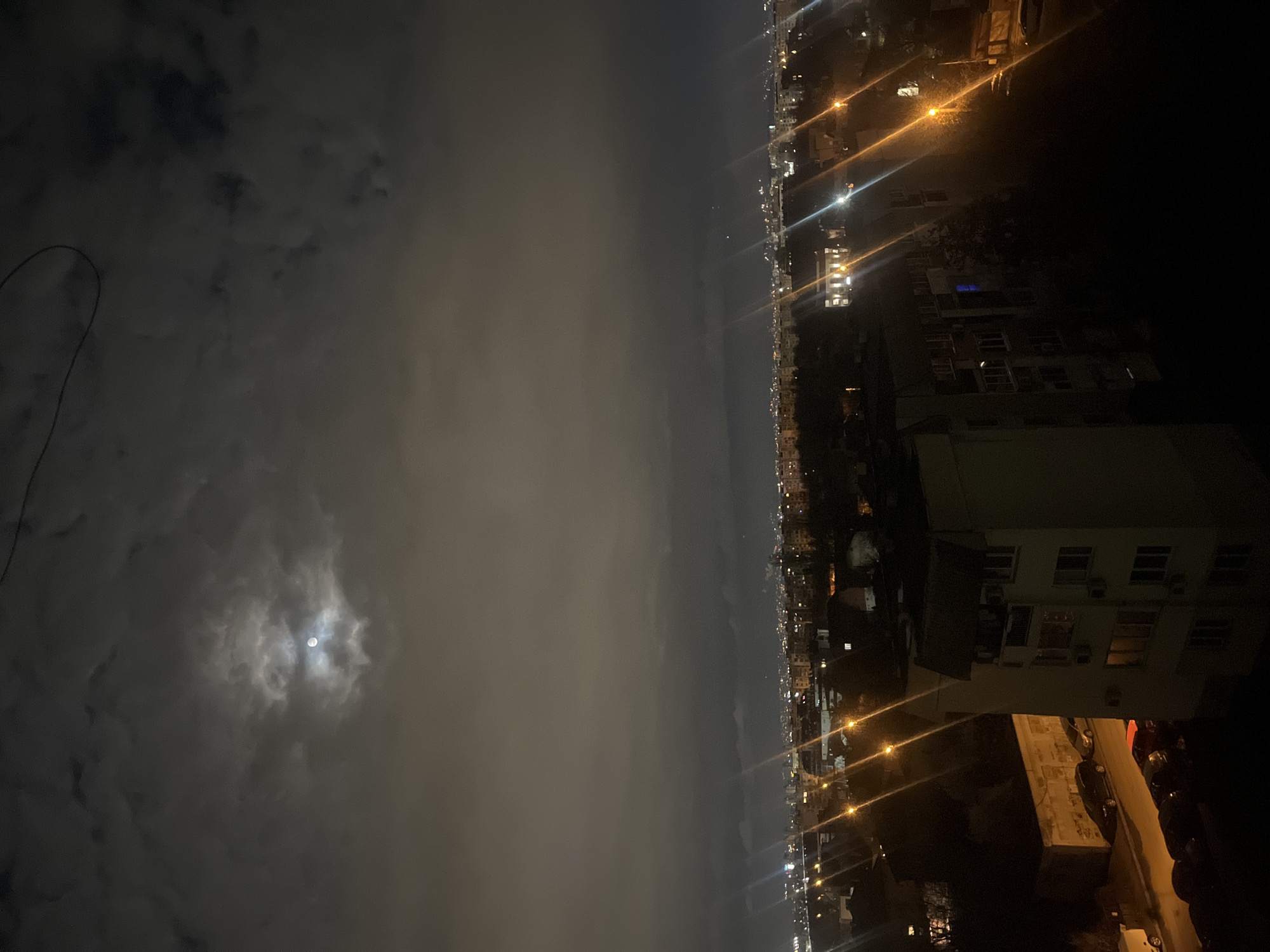 A night cityscape, possibly in Sofia, Bulgaria, shows a clouded sky with the moon visible on the left. The right side shows buildings and streetlights, indicating urban development. The image suggests an elevated viewpoint overlooking the city at night.