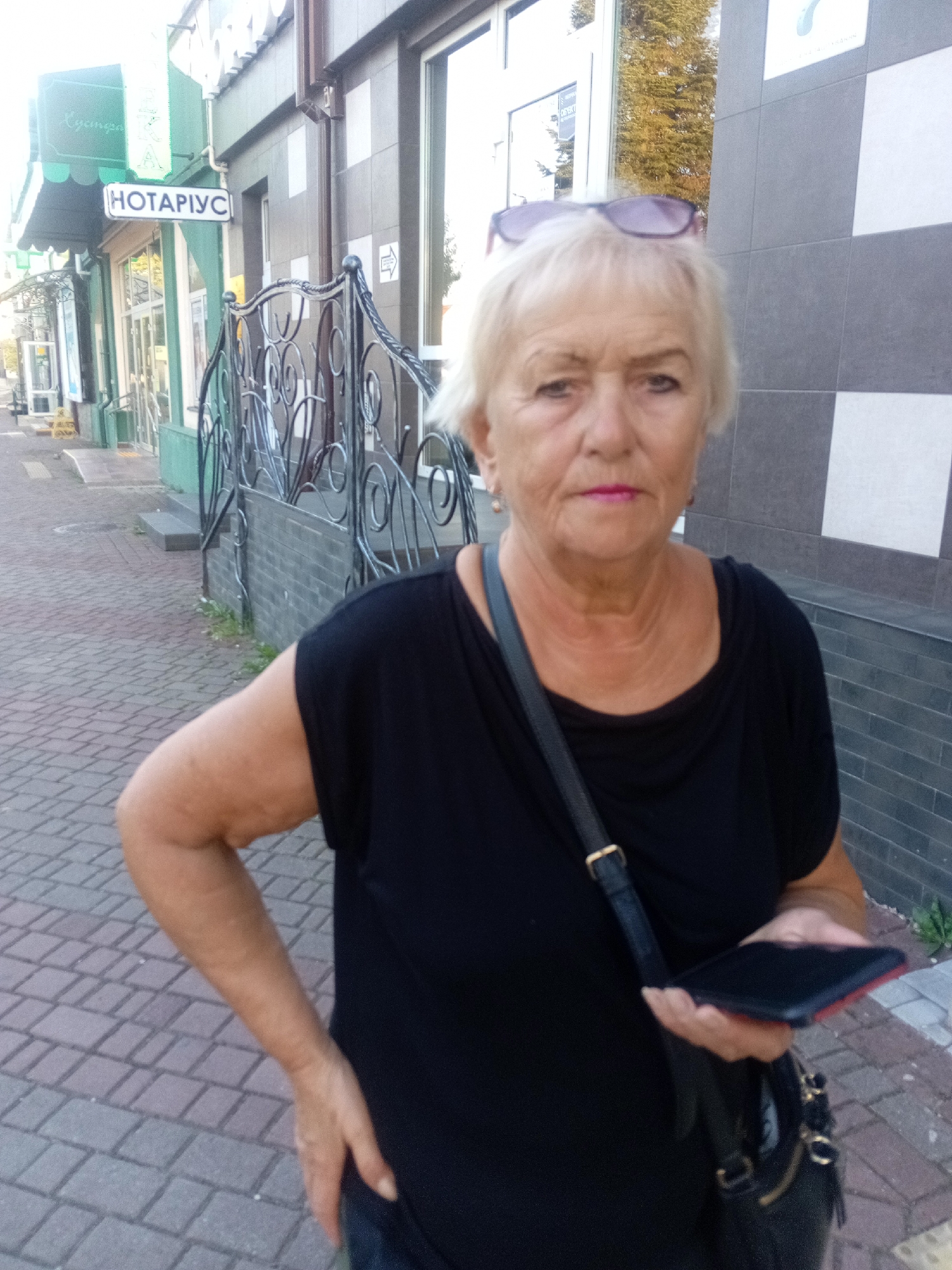 A blonde, middle-aged woman with sunglasses perched on her head and wearing a black V-neck top and jeans stands on a brick sidewalk in front of a building in Tiachiv, Ukraine. She is holding a black smartphone in her right hand. The building behind her has a dark brick lower section and lighter tiled upper sections. A wrought iron fence with decorative scrollwork is in front of the building. To the left of the woman, a green awning extends over a storefront with a sign that reads "НОТАРІУС" (NOTARY). The sidewalk is paved with red and gray bricks laid in a herringbone pattern.