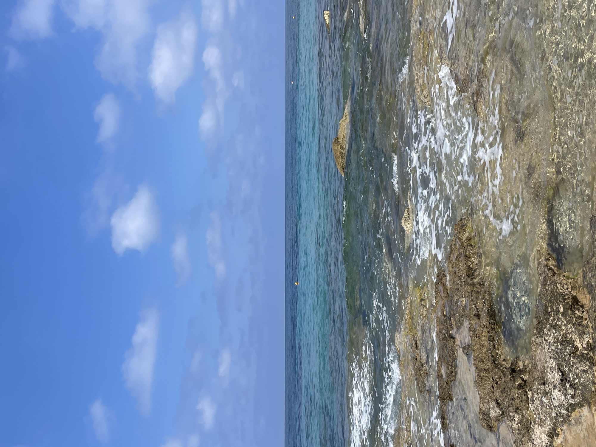 A low-angle, eye-level shot captures a tranquil coastal scene on a clear day, likely in Paphos, Cyprus. The horizon is divided into three horizontal bands: a vibrant blue sky with scattered, puffy white clouds in the upper third; a calm, cerulean sea with a darker blue line on the horizon in the middle third; and the foreground is occupied by shallow, clear water lapping over rocky terrain.
The rocky shore is composed of large, uneven, and weathered stones, some submerged in the water and others exposed. The water over the rocks is transparent, revealing the seabed beneath. Gentle waves create white foam as they break against the rocks and recede.
The sea itself exhibits varying shades of blue and turquoise, indicating depth and clarity. In the middle distance, a few small, yellow buoys are visible, suggesting a designated swimming area or marker. The overall impression is one of natural beauty and serenity. There are no people or animals present in the frame.