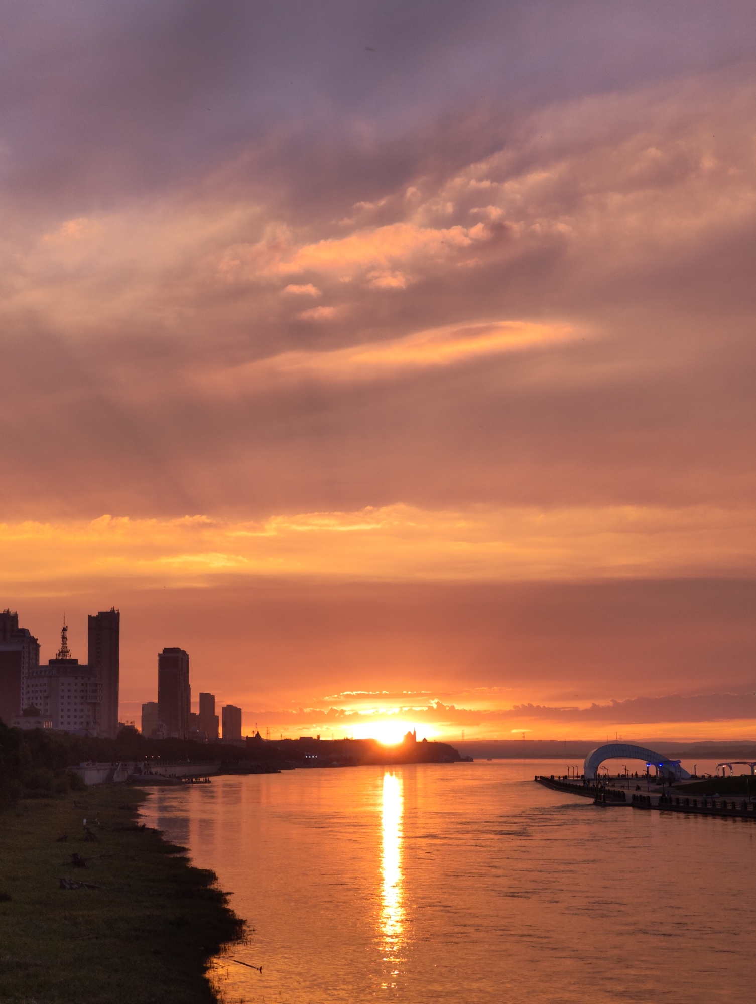 This is a vertical, eye-level, medium shot of a riverfront cityscape at sunset. The sky is a dramatic display of oranges, pinks, and purples, with clouds catching the setting sun's light. The sun itself is partially obscured by clouds and is seen as a bright, radiant orb near the horizon, casting a shimmering reflection on the water.
In the foreground, a wide river occupies the bottom half of the frame, its surface reflecting the vibrant colors of the sky. The left bank of the river is lined with green grass and some scattered debris. Further inland, a silhouette of a city skyline is visible, featuring several tall, modern buildings. One building has a distinctive antenna on top.
On the right side of the frame, a modern architectural structure, resembling a bridge or archway with lights, juts out into the river from a pier. Several small figures of people are visible on the pier, appearing as dark silhouettes against the bright water and sky. The overall atmosphere is serene and picturesque, typical of a sunset over a developed riverfront. The location is identified as 幸福乡, China.