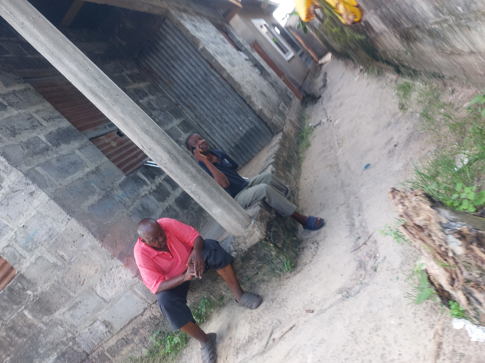 This media file captures an outdoor scene in what appears to be a residential or semi-urban area in Dar es-Salaam, Tanzania, with a distinctive tilted perspective. Two men are the primary subjects, seated near a building constructed from concrete blocks and corrugated metal.
In the foreground and to the left, an older man with a bald head wears a short-sleeved red polo shirt and dark shorts or cropped trousers, paired with grey slip-on sandals. He is seated directly on the dirt ground, leaning against a low concrete structure, with his hands clasped together or resting in front of him. His gaze appears directed downwards or slightly forward.
Partially obscured by a diagonal light-colored wooden or concrete pillar, another man is visible sitting on a raised concrete ledge behind the first man. He is wearing a dark blue or black short-sleeved shirt and light-colored trousers, with blue sandals. This man is actively engaged in a phone call, holding a mobile phone to his right ear.
The immediate setting includes a sandy or dirt path interspersed with patches of green grass and small pieces of debris. To the far right, a large piece of weathered wood or a log is visible on the ground. The background features more of the informal building structures with corrugated metal panels and simple windows. The lighting suggests it is daytime, possibly late morning or afternoon, with no harsh shadows indicating direct midday sun. There is no visible text in the image. The overall impression is one of everyday life, with the men casually resting or socializing.