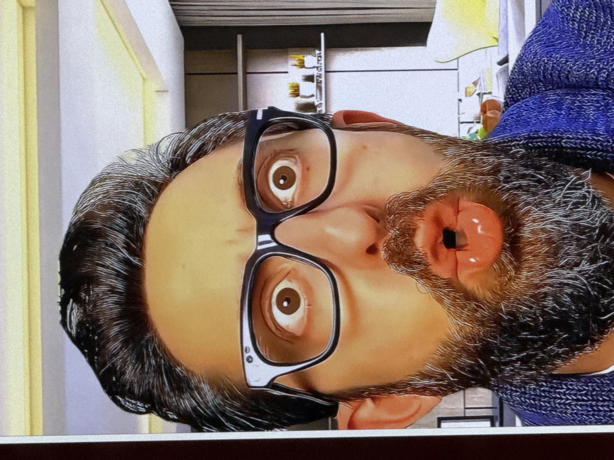 The image shows a close-up, front-facing view of a person's face with exaggerated features. The person has dark, styled hair, is wearing thick-rimmed black glasses, and has a full beard. Their eyes are wide open, and their mouth is puckered as if about to kiss or whistle. The background appears to be an indoor setting, possibly a living room or study, with shelves and furniture visible. There is no information in the image that suggests a location in Cahul, Moldova.