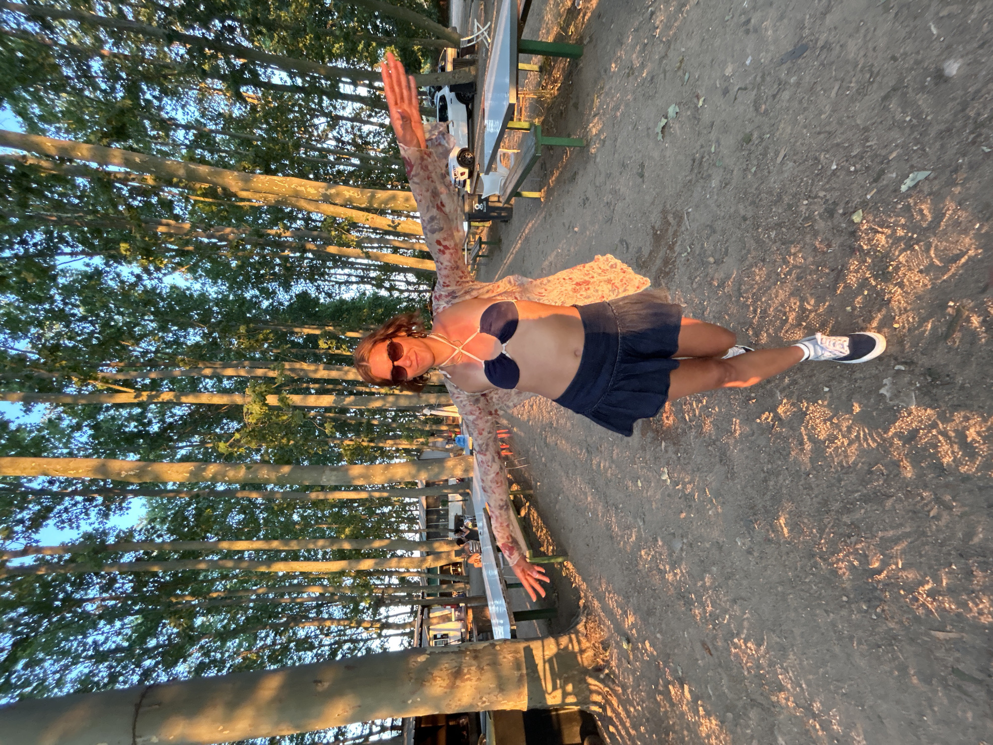The image shows a young woman with brown hair and sunglasses standing in an outdoor area with a sandy ground, surrounded by tall trees. She is wearing a dark blue bikini top, a dark blue skirt, and a floral-patterned open cardigan. Her arms are extended outwards. In the background, there are picnic tables, benches, some buildings, and parked cars. The sun is low, casting long shadows and a warm light on the scene, suggesting it is either sunrise or sunset. The location appears to be a campground or an outdoor dining area, possibly near a beach or a natural park, and the context suggests it is a recreational area.