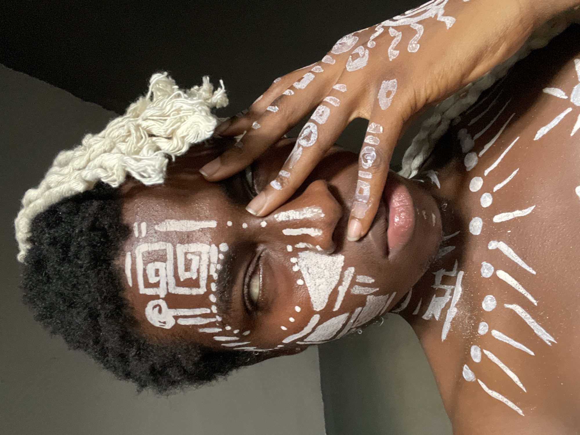 A person with dark skin and a head of black curly hair, adorned with white paint markings on their face and body, is seen from a close-up, tilted perspective. The person’s right hand, also decorated with white markings, is positioned over their face with fingers touching the forehead and cheek. They appear to be wearing a rope or yarn-like material on their head, partially covering their hair. The background is a combination of dark and light areas, suggesting an indoor setting. The body paint markings resemble tribal or cultural designs. The image does not contain any information to verify or determine the city of Onitsha, Nigeria.