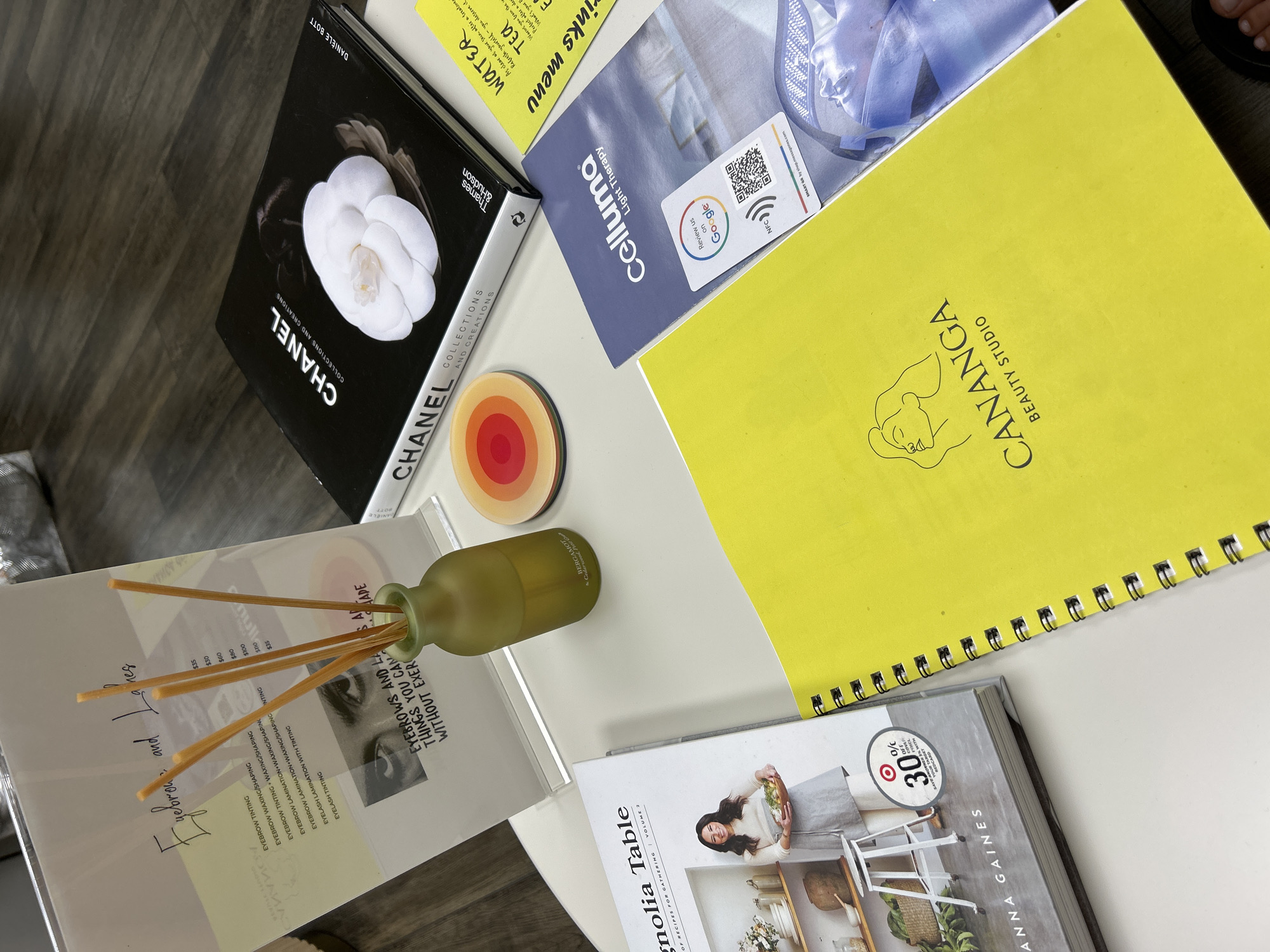 The image shows a white round table with several items on it, suggesting a beauty studio or a similar establishment. Among the objects are a book titled "CHANEL COLLECTIONS AND CREATIONS," a cookbook titled "Magnolia Table" by Joanna Gaines, a yellow notebook with "CANANGA BEAUTY STUDIO" printed on it, and a brochure for "Celluma Light Therapy." There's also a small glass vase with reeds, a coaster with a colorful design, and a sign listing eyebrow and lash services with prices. A part of a person's foot is visible at the bottom right of the image. The overall setting appears to be indoors, with a wooden floor visible in the background. The items suggest a contemporary and perhaps upscale environment related to beauty and lifestyle. There is no specific indication of the city of Hallandale Beach, United States of America.